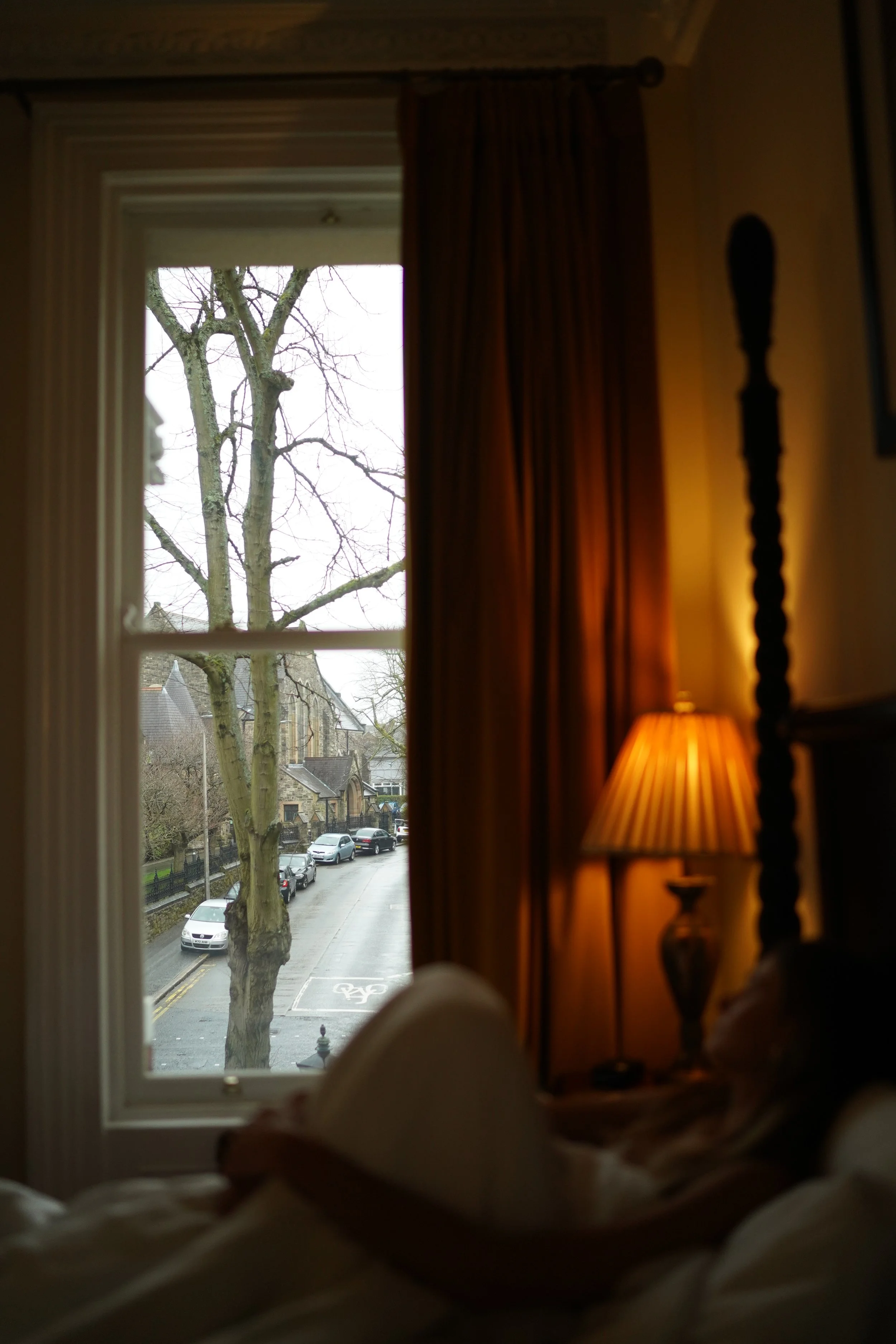 View from a dimly lit room showing a window with a street scene outside; includes a tree, buildings, and parked cars. Inside, there's a person lying on a bed and a lit lamp on a side table.