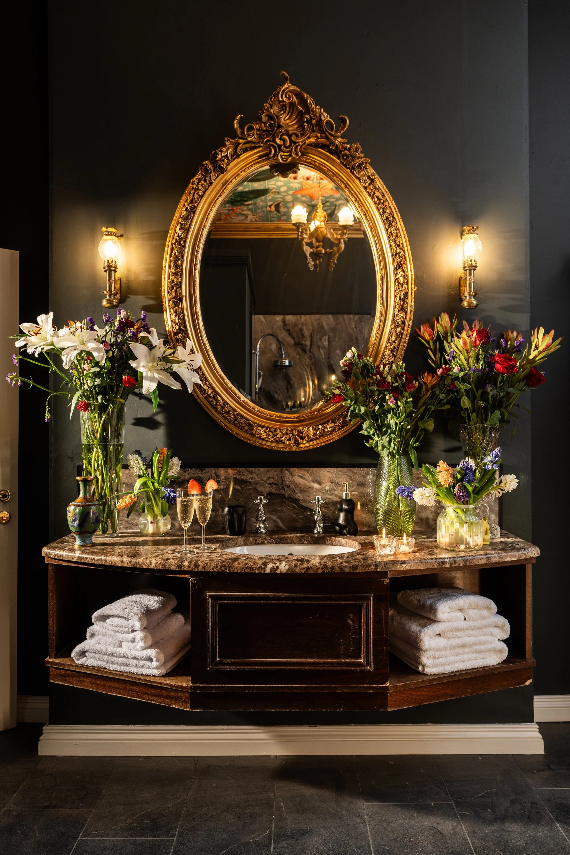 Elegant bathroom vanity with a marble countertop, ornate gold-framed mirror, and floral arrangements. Glasses of champagne, candles, and neatly stacked white towels complete the luxurious setting.