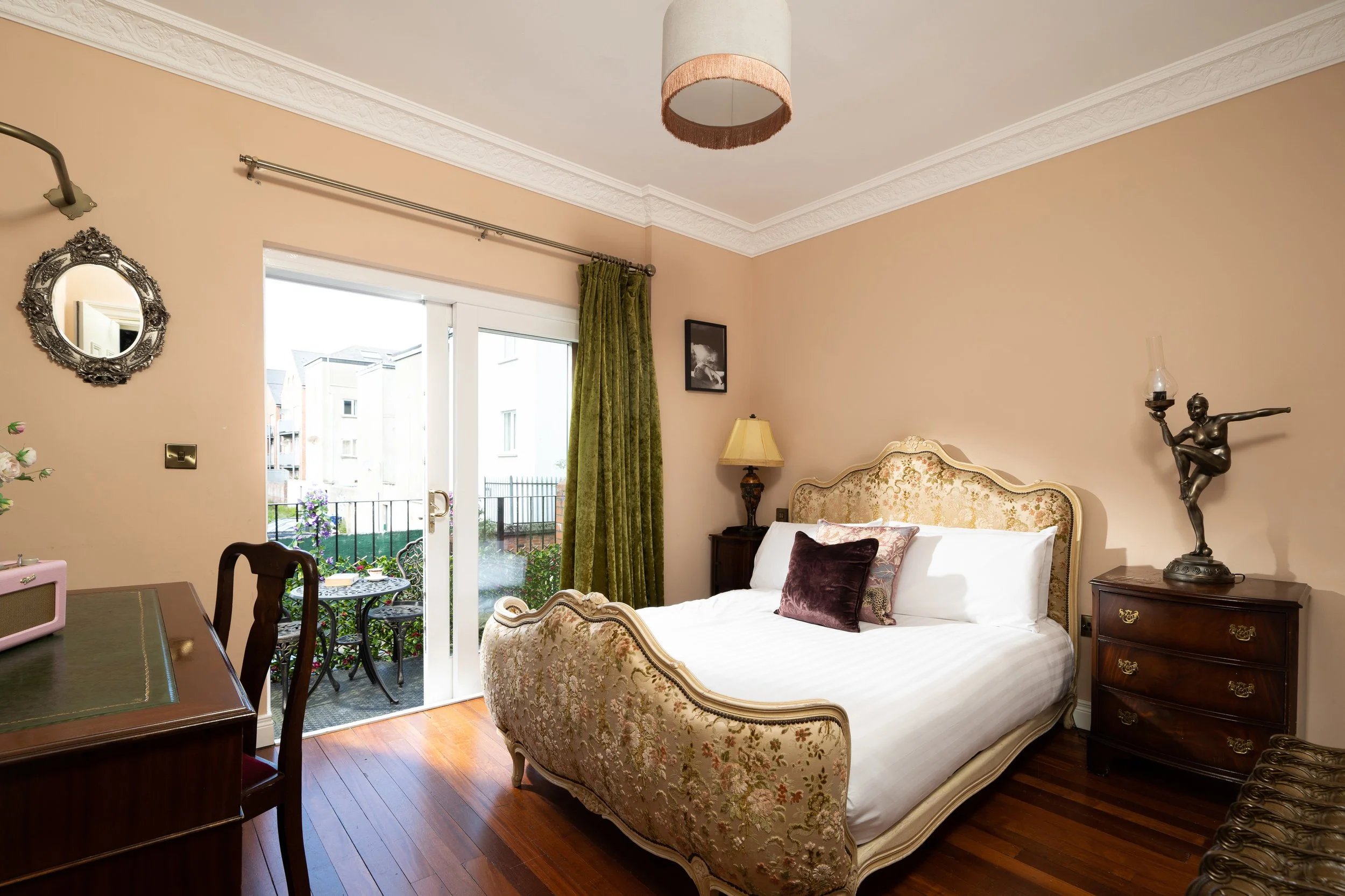 Elegant bedroom with a vintage-style bed, ornate mirror, and wooden furniture with a sculpture lamp. A door opens to an outdoor patio with a small table and chairs.