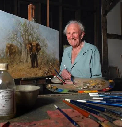 An elderly woman standing at an art easel, painting a landscape scene featuring two elephants in a dry, grassy area with trees. She is holding a paintbrush and smiling, with a palette of colors in front of her.