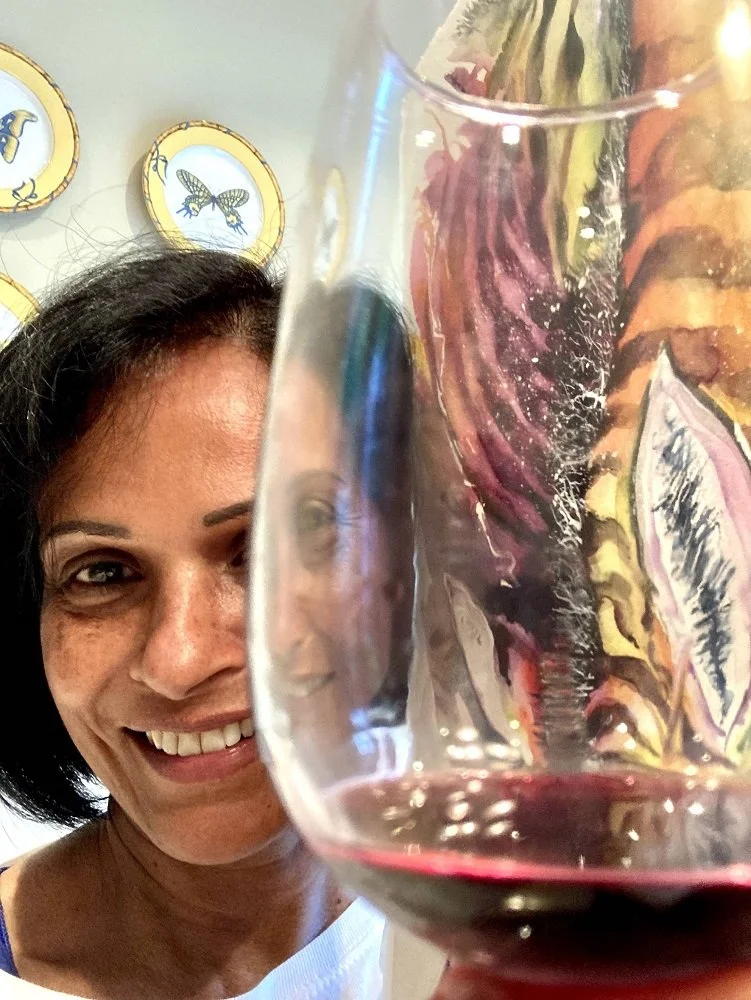 A woman smiling and holding a glass of red wine in front of her face, with colorful artwork and bee-themed plates in the background.