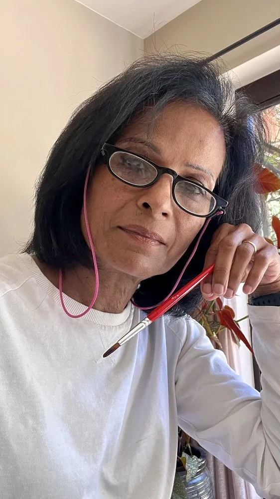 Woman with black hair, glasses, and a white shirt holding a calligraphy brush near her face, indoors near a window with plants in the background.