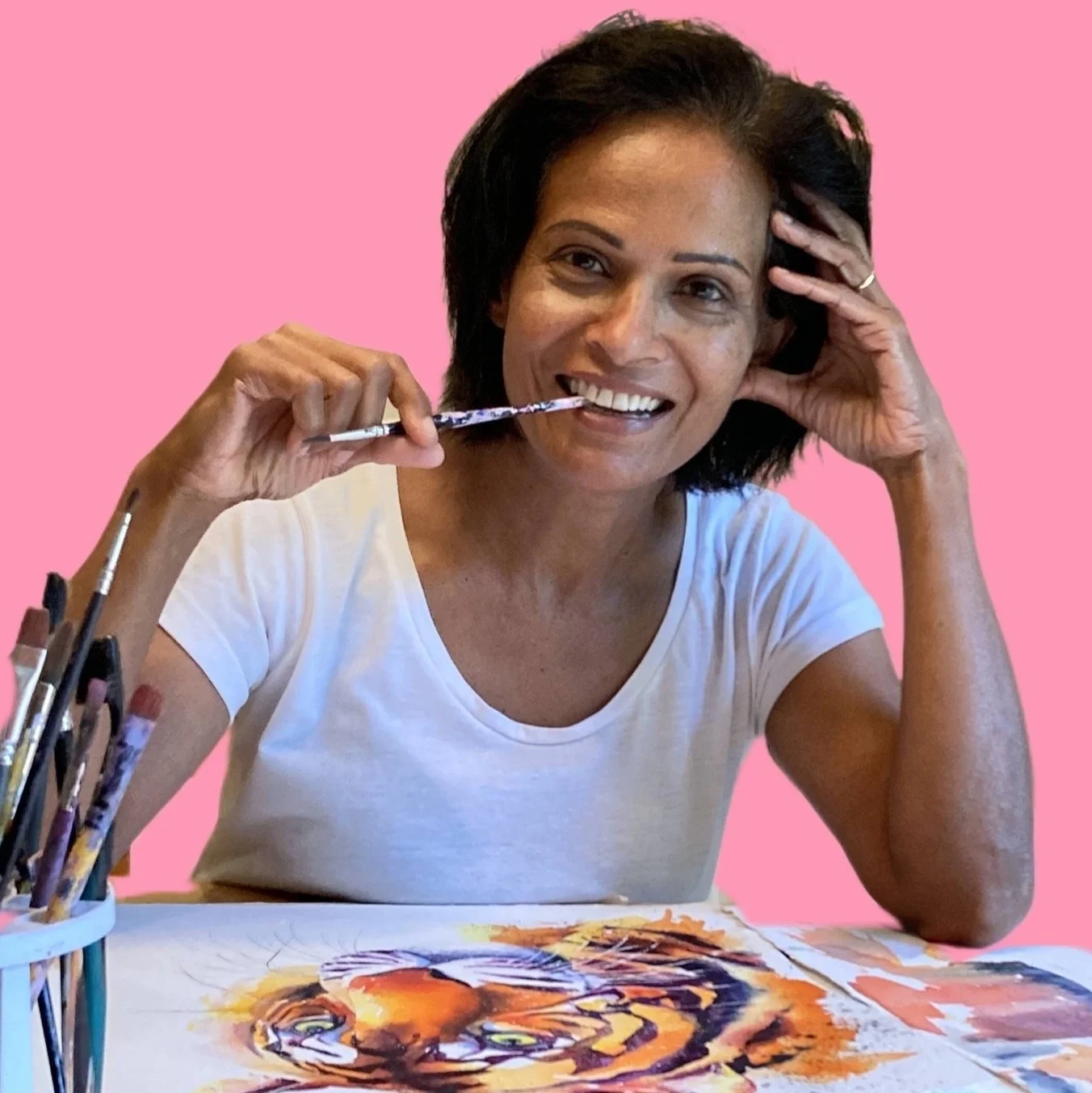 A woman with dark hair smiling, holding a paintbrush near her mouth, sitting at a table with a colorful watercolor painting of a tiger, against a pink background.
