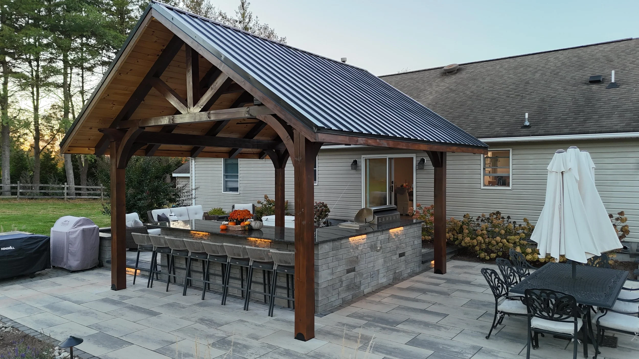 Backyard patio with a covered outdoor kitchen and bar area, featuring a metal roof, stone counter with barstools, a grill, outdoor dining table with chairs and umbrella, surrounded by plants and a house with vinyl siding.