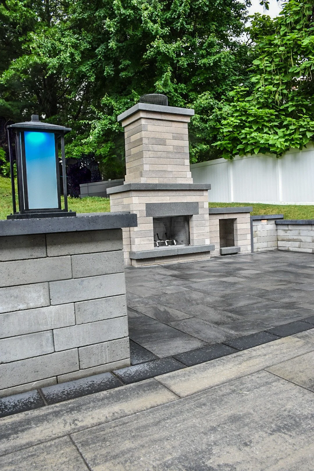 An outdoor patio area with a brick fireplace, a stone flooring, and a white fence in the background, surrounded by green trees.
