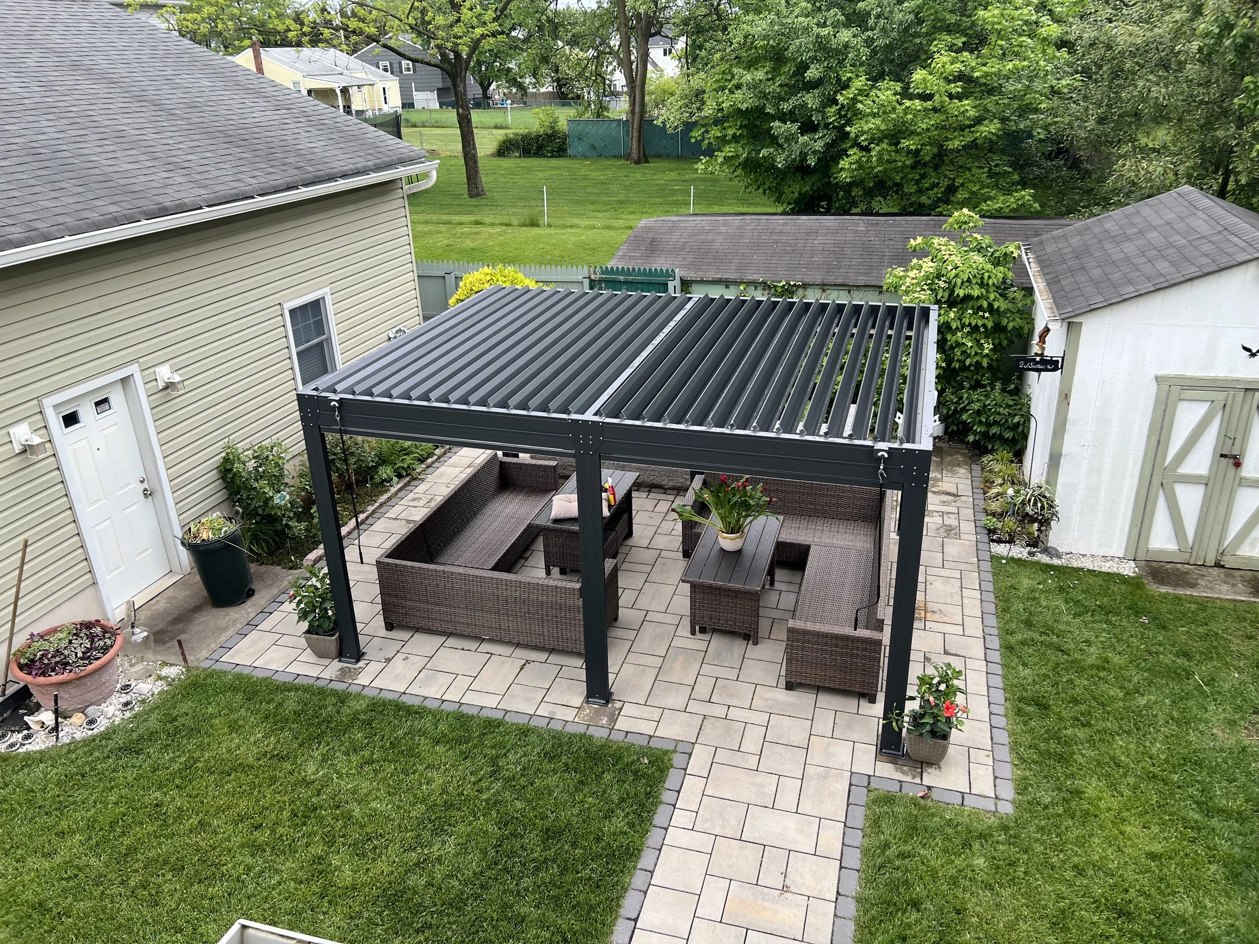 A backyard patio area with a black metal pergola, outdoor wicker furniture, potted plants, and a well-maintained lawn surrounded by trees and neighboring houses.