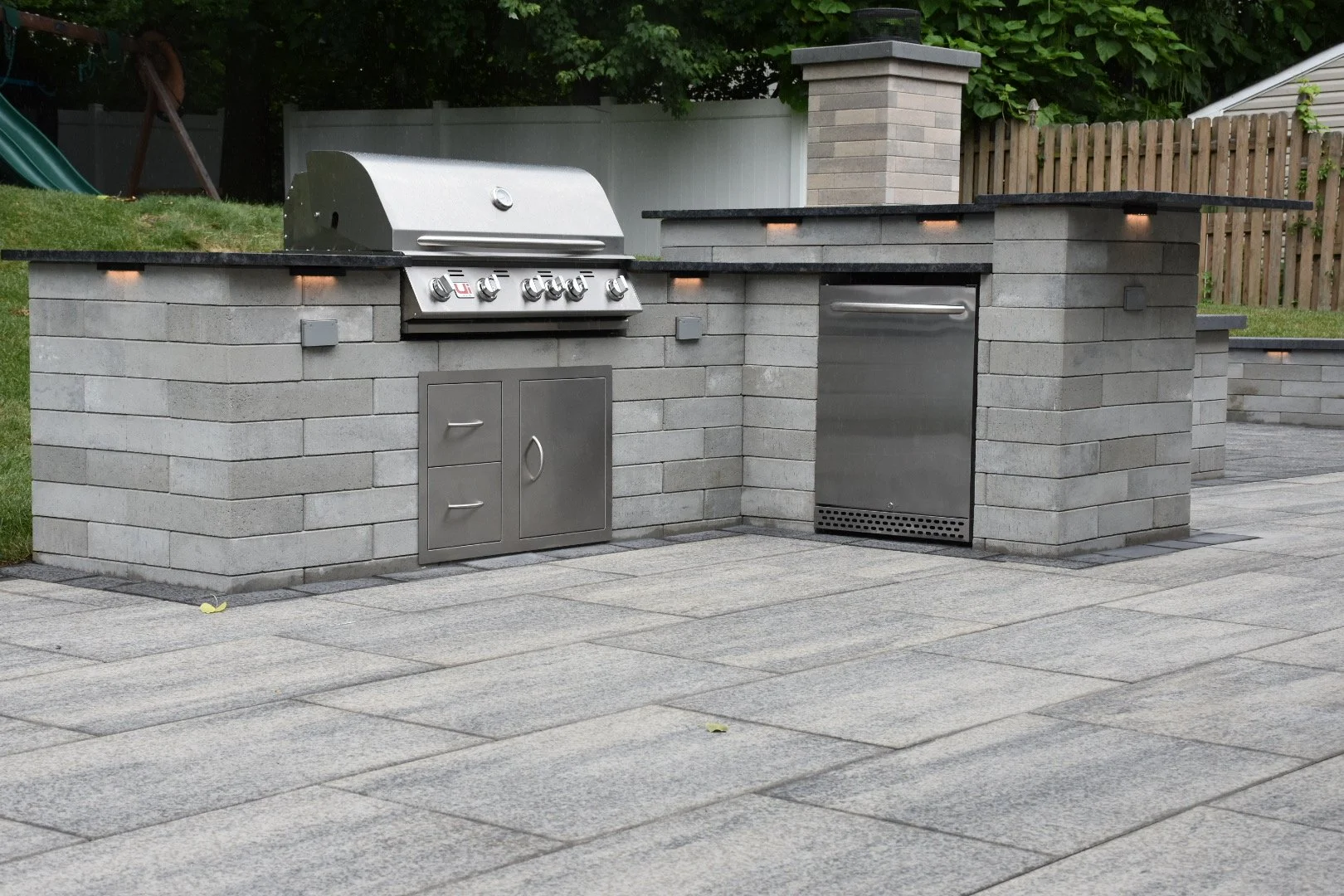 Outdoor kitchen with built-in grill, stainless steel refrigerator, and storage cabinets, set on a stone-paved patio with a brick and stone wall background.