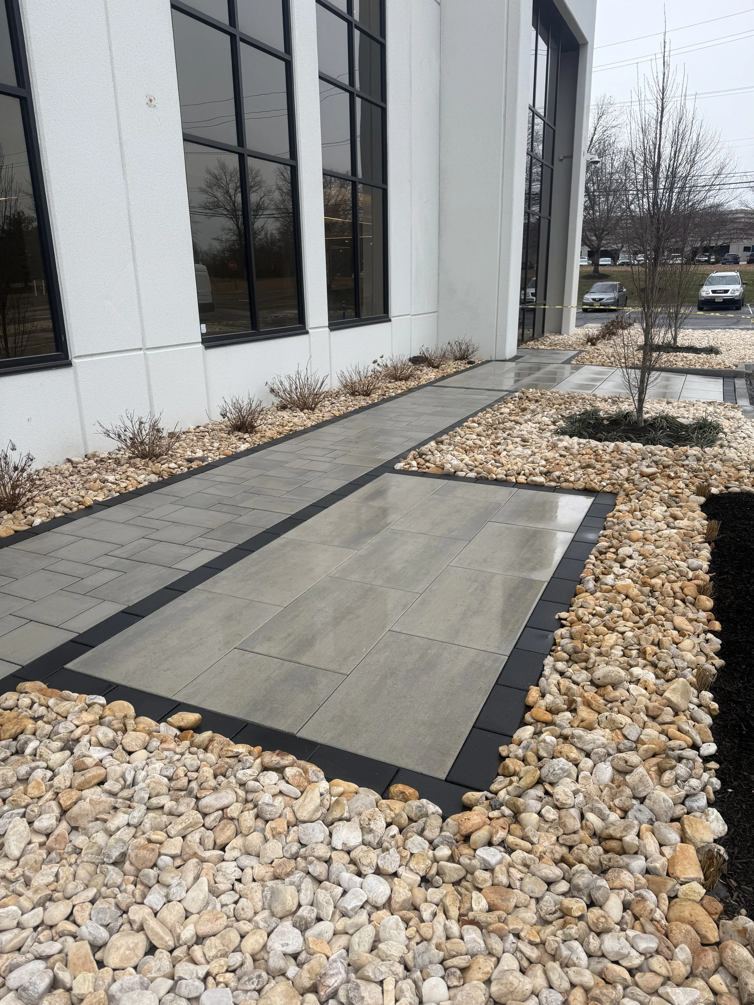 Concrete walkway with decorative border, surrounded by landscaped areas with rocks, small bushes, and trees outside a building with large windows.