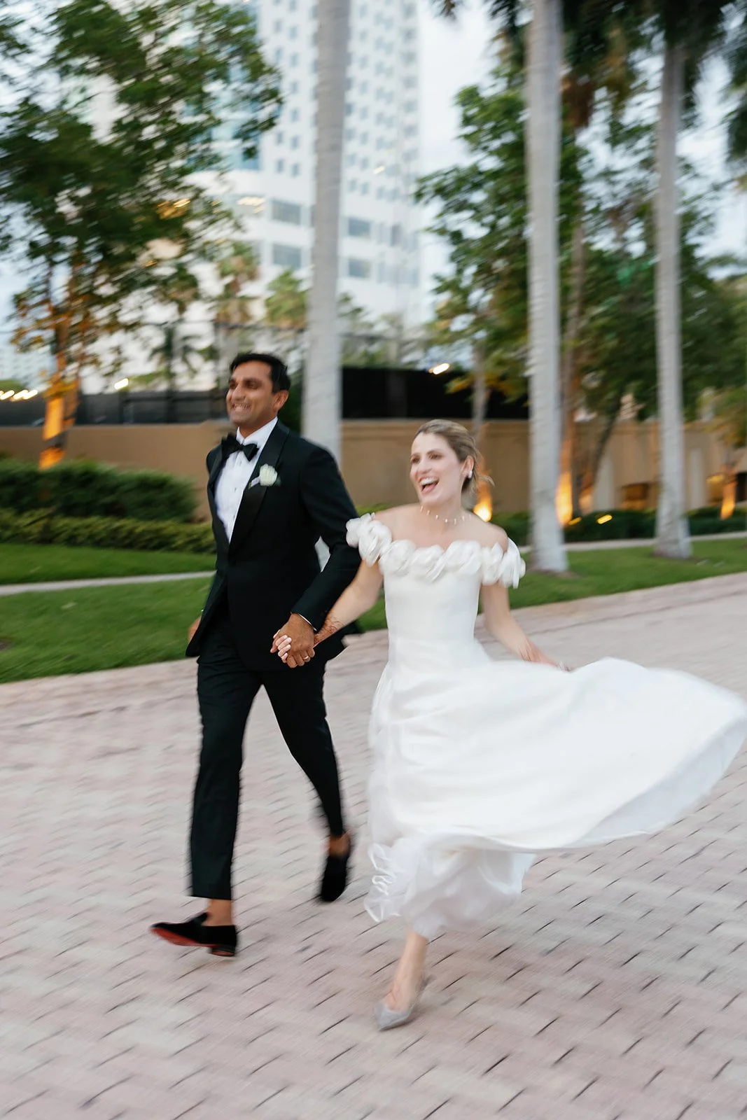 A bride and groom are running down a walkway outdoors, holding hands. 