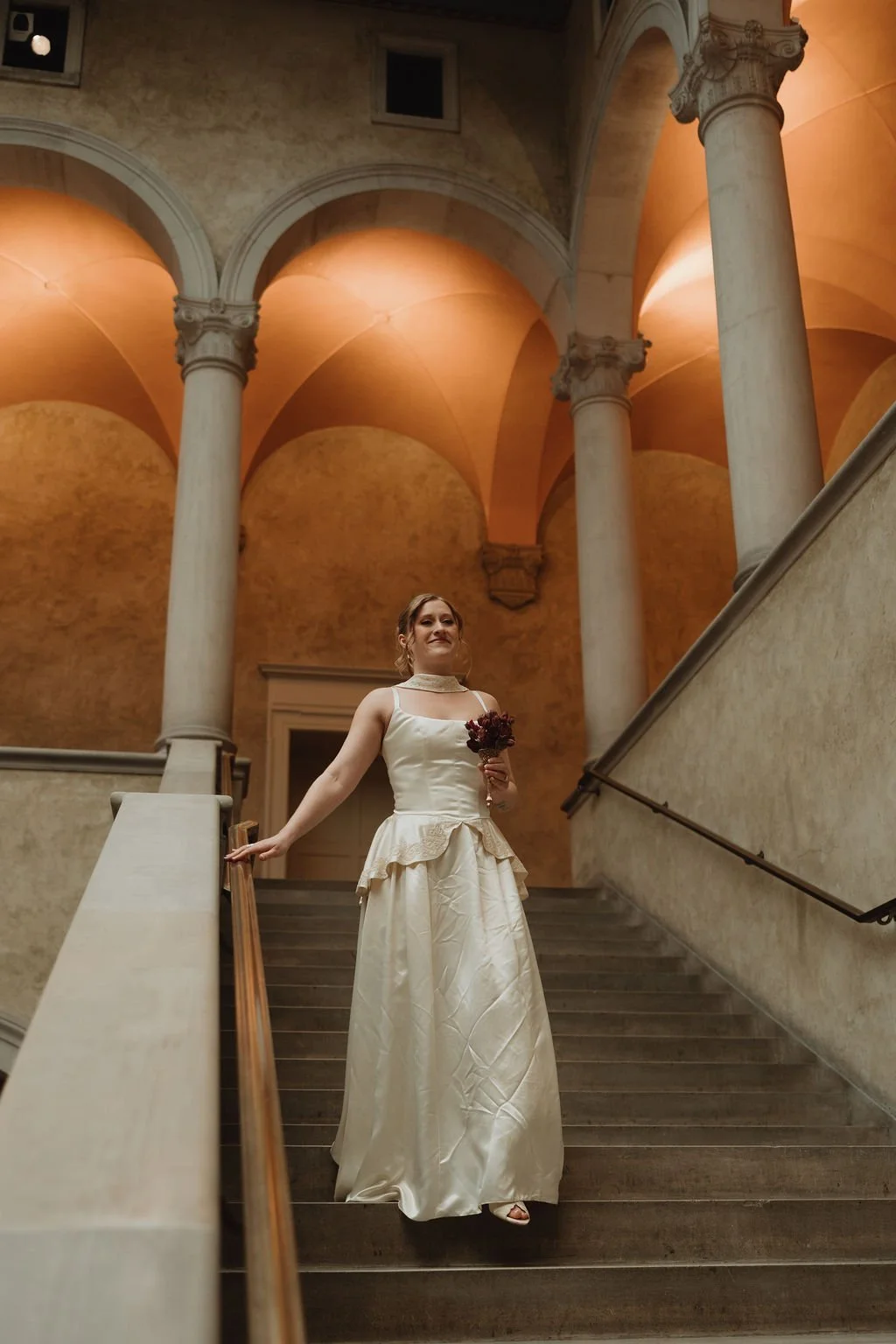 A woman in a wedding dress holding a bouquet of flowers standing on a staircase inside a grand building with marble columns and arched ceilings.