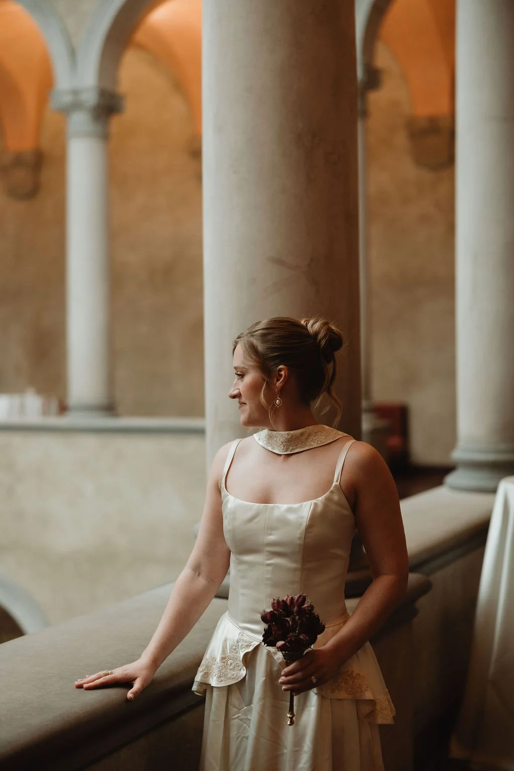 A bride in a white wedding dress holding a small bouquet of dark red flowers, standing in an elegant venue with tall columns and arched ceilings.