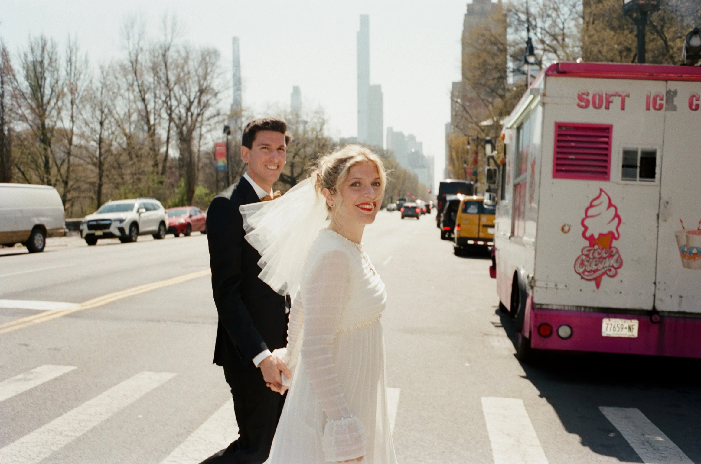 A bride wearing a wedding veil and holding hands with her groom are crossing an NYC street with an ice cream truck parked on the side.