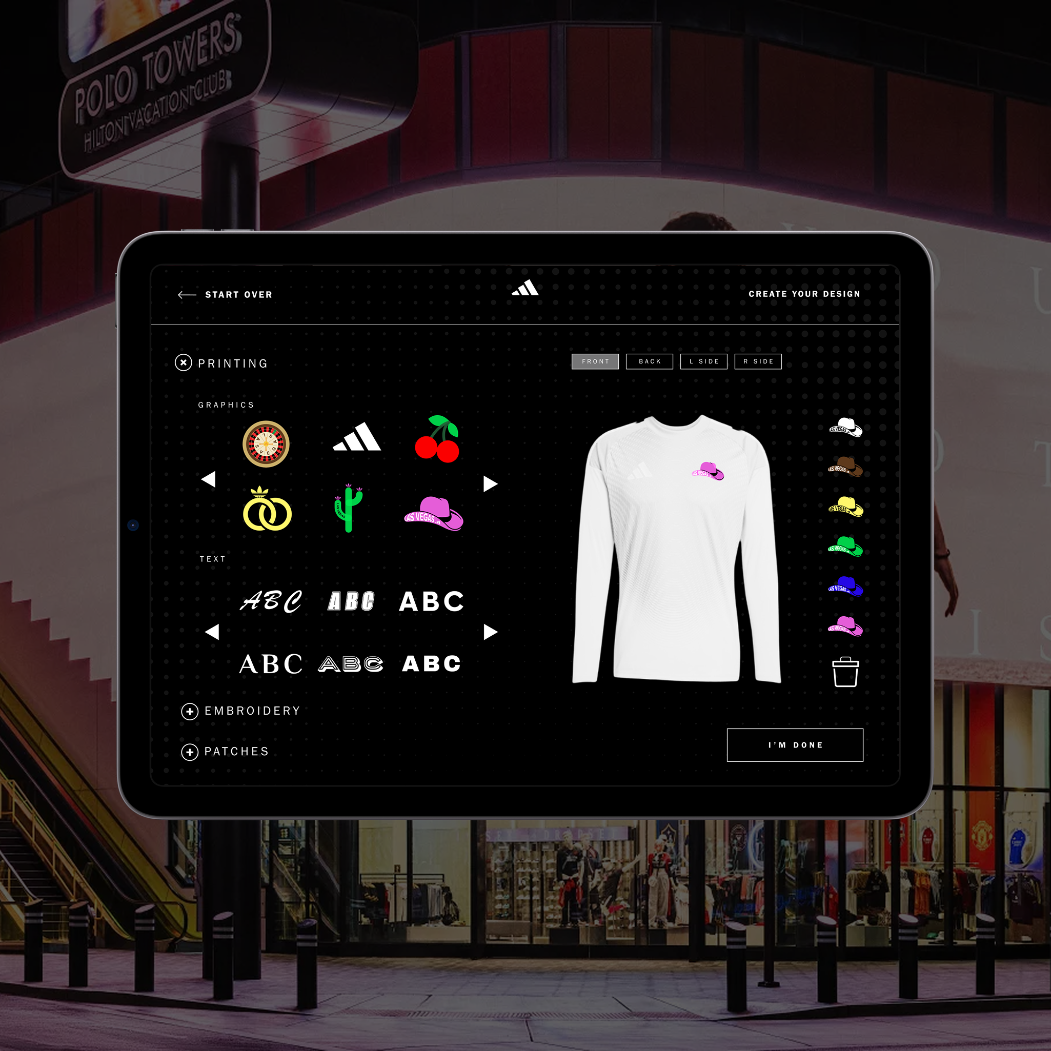 A tablet screen showing a digital t-shirt customization interface with various design options, including graphics, text, embroidery, and patches, set against a background of a shopping mall exterior at night.