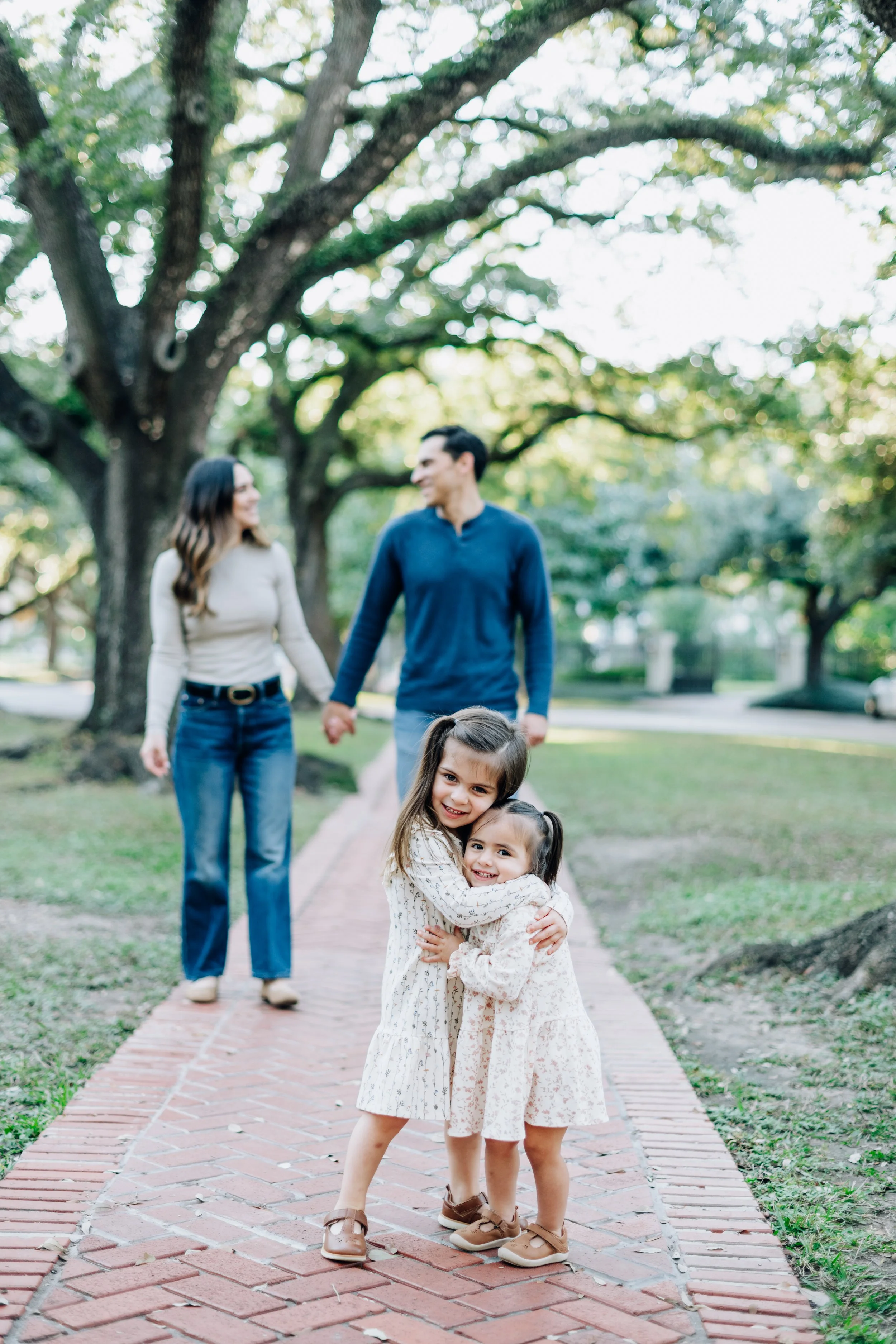 Two young girls hug and smile at the camera on a brick path in a park, with a couple holding hands and smiling in the background beneath a large tree.
