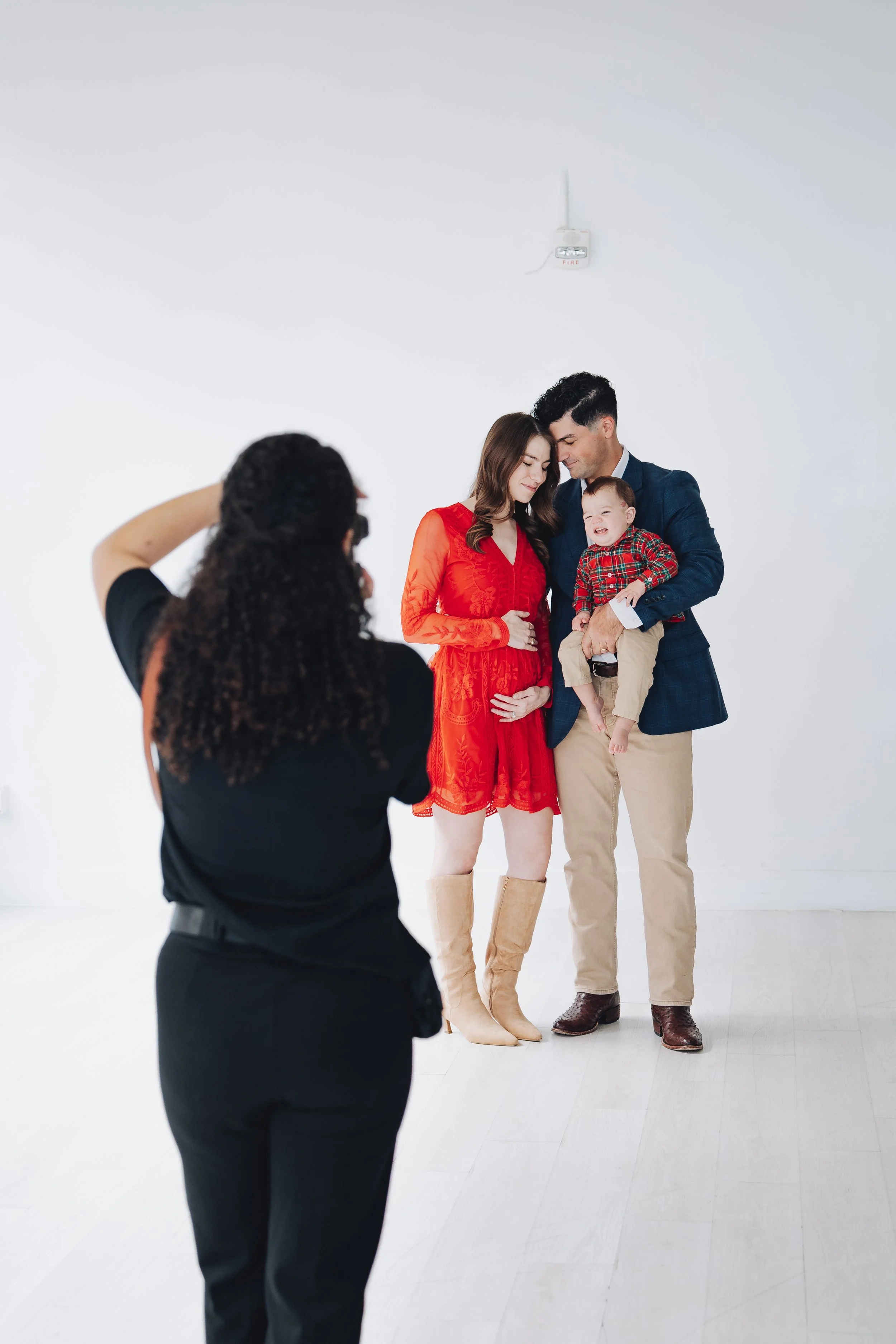 A family photo session with a woman in a red dress and beige boots, a man in a blue blazer holding a young boy in a plaid shirt, and a photographer taking their picture against a plain white wall.