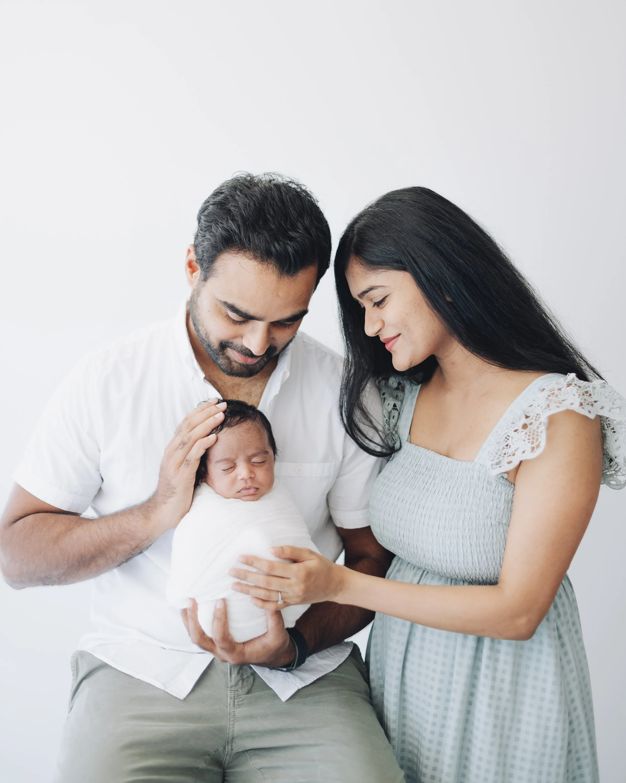 A family consisting of a man, woman, and newborn baby, with the man and woman gently holding the baby and smiling at each other.