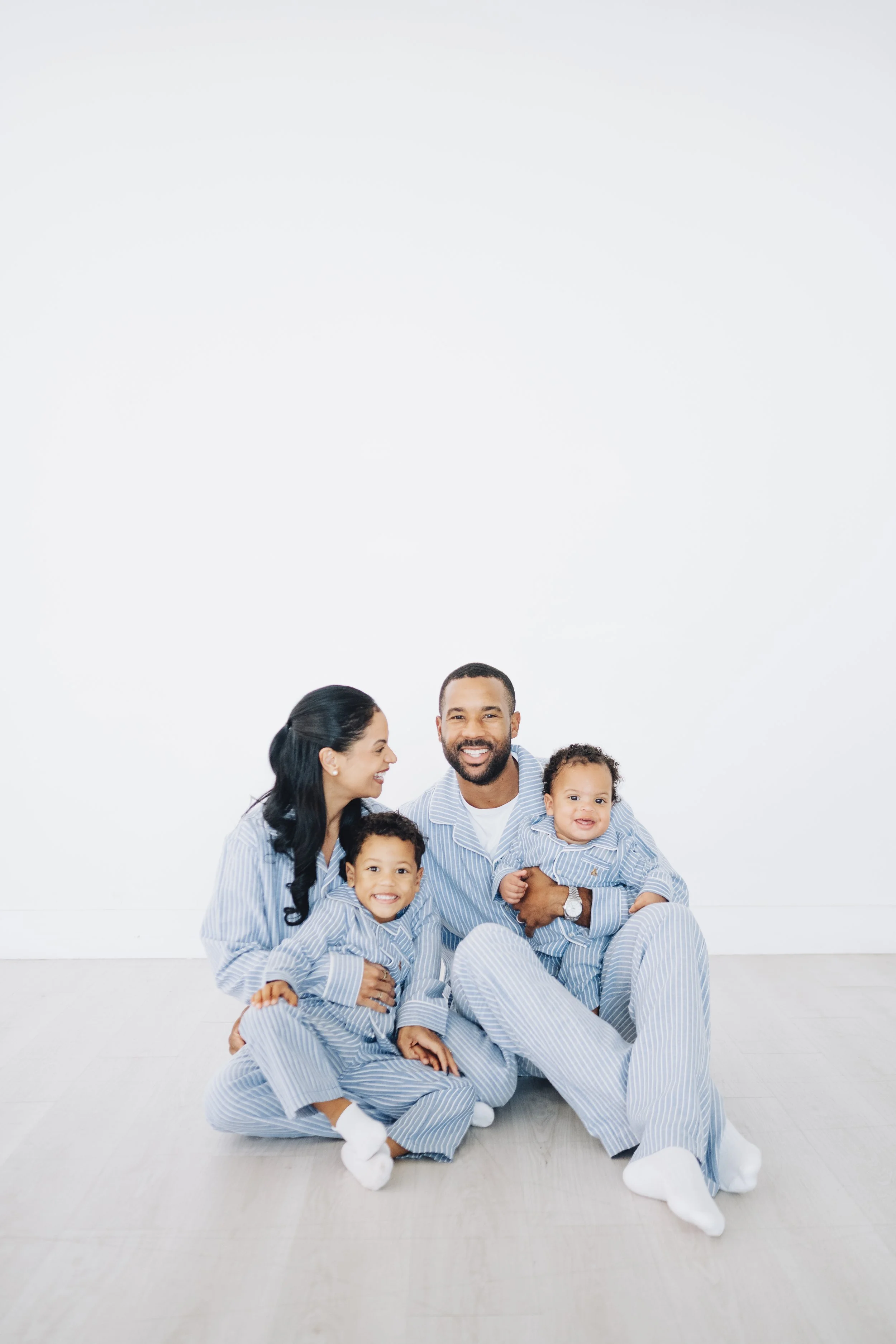 Family of four sitting on the floor in pajamas, smiling and embracing each other in a bright, white room.