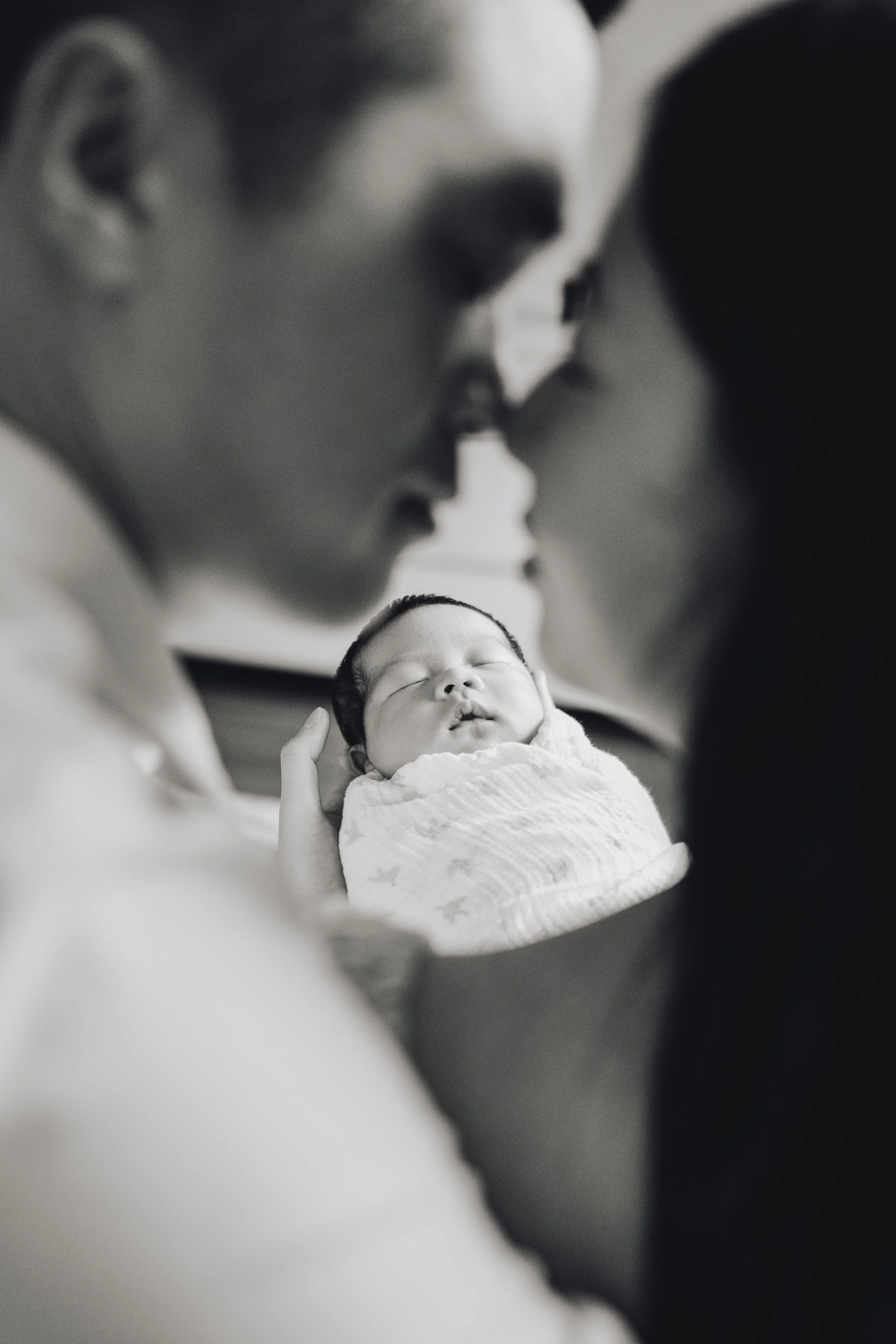 A black-and-white photo of two adults, likely the parents, gently touching their foreheads together, holding their newborn baby between them. The baby is peacefully sleeping and wrapped in a blanket.