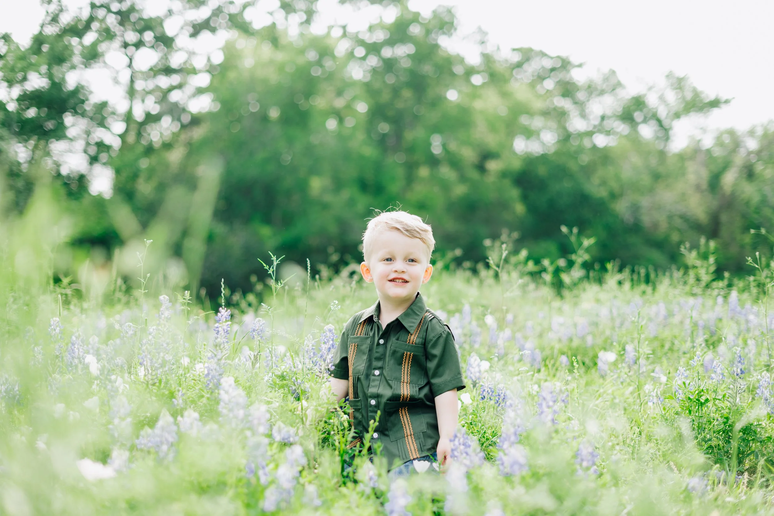 A young boy with blonde hair in a dark green shirt standing in a field of purple and white flowers with trees in the background.