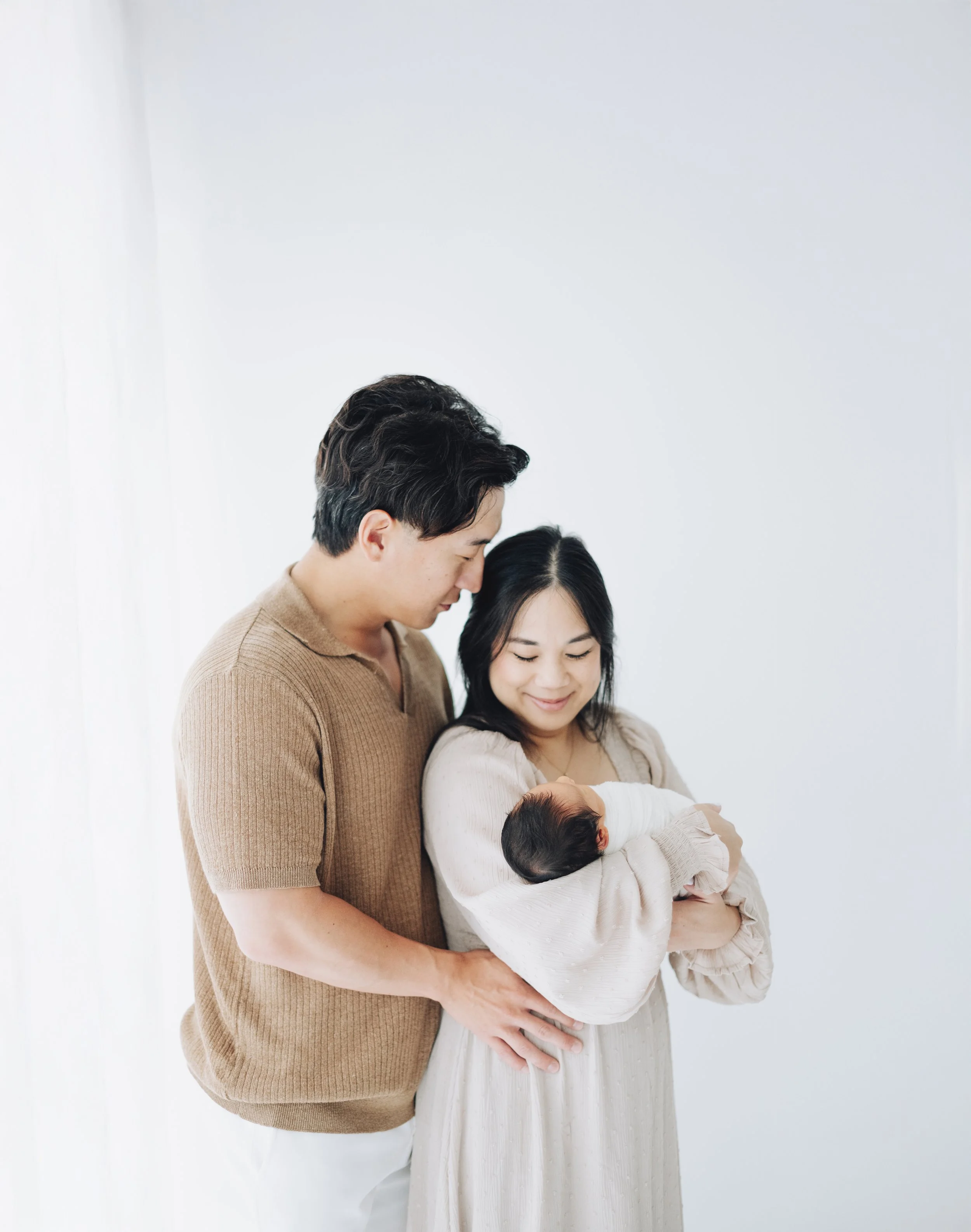 An Asian couple holding their newborn baby indoors against a plain light-colored wall. The mother is smiling while cradling the baby, and the father is gently holding her around the waist, both looking at the baby lovingly.