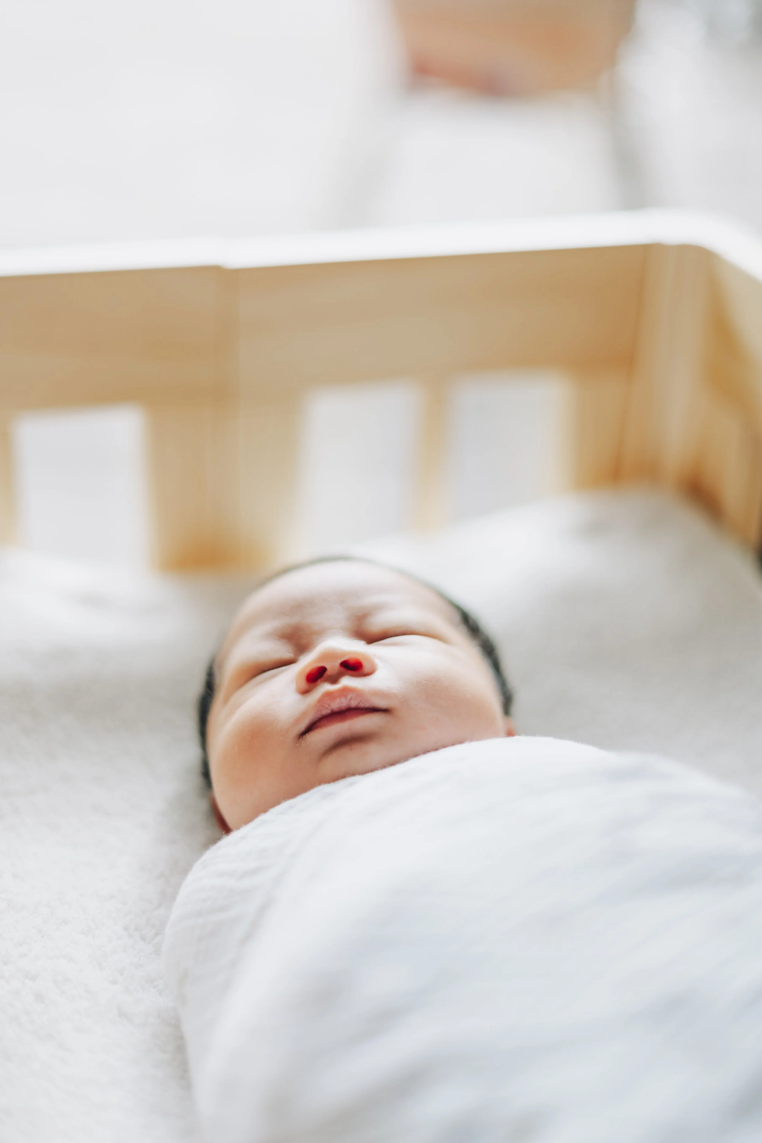 Close-up of a newborn baby sleeping peacefully in a wooden crib, wrapped in a white blanket.