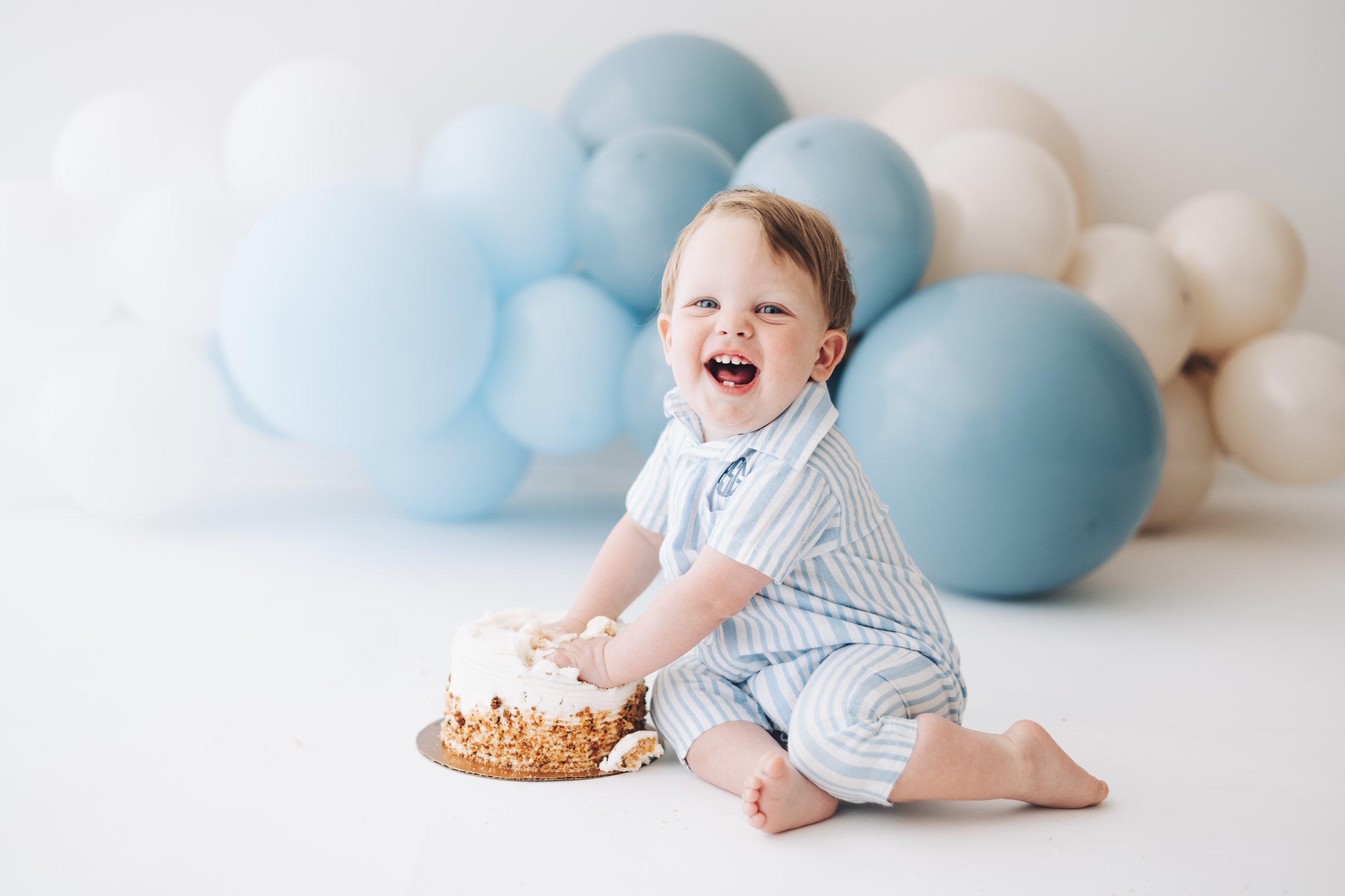 A happy young child in striped pajamas celebrating with a cake, surrounded by blue and white balloons.