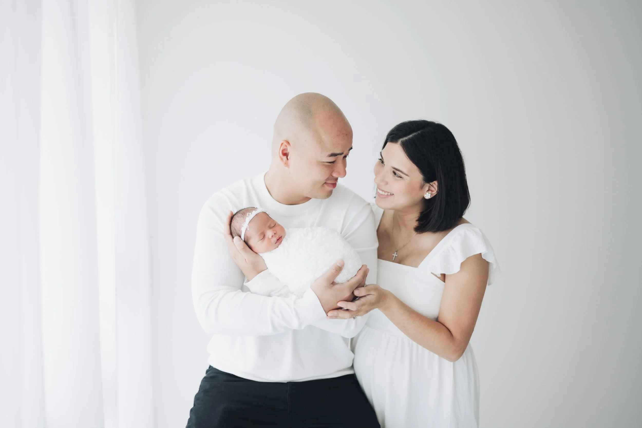 A happy family of three, including a pregnant woman, a man, and a newborn baby, dressed in white in a bright room.
