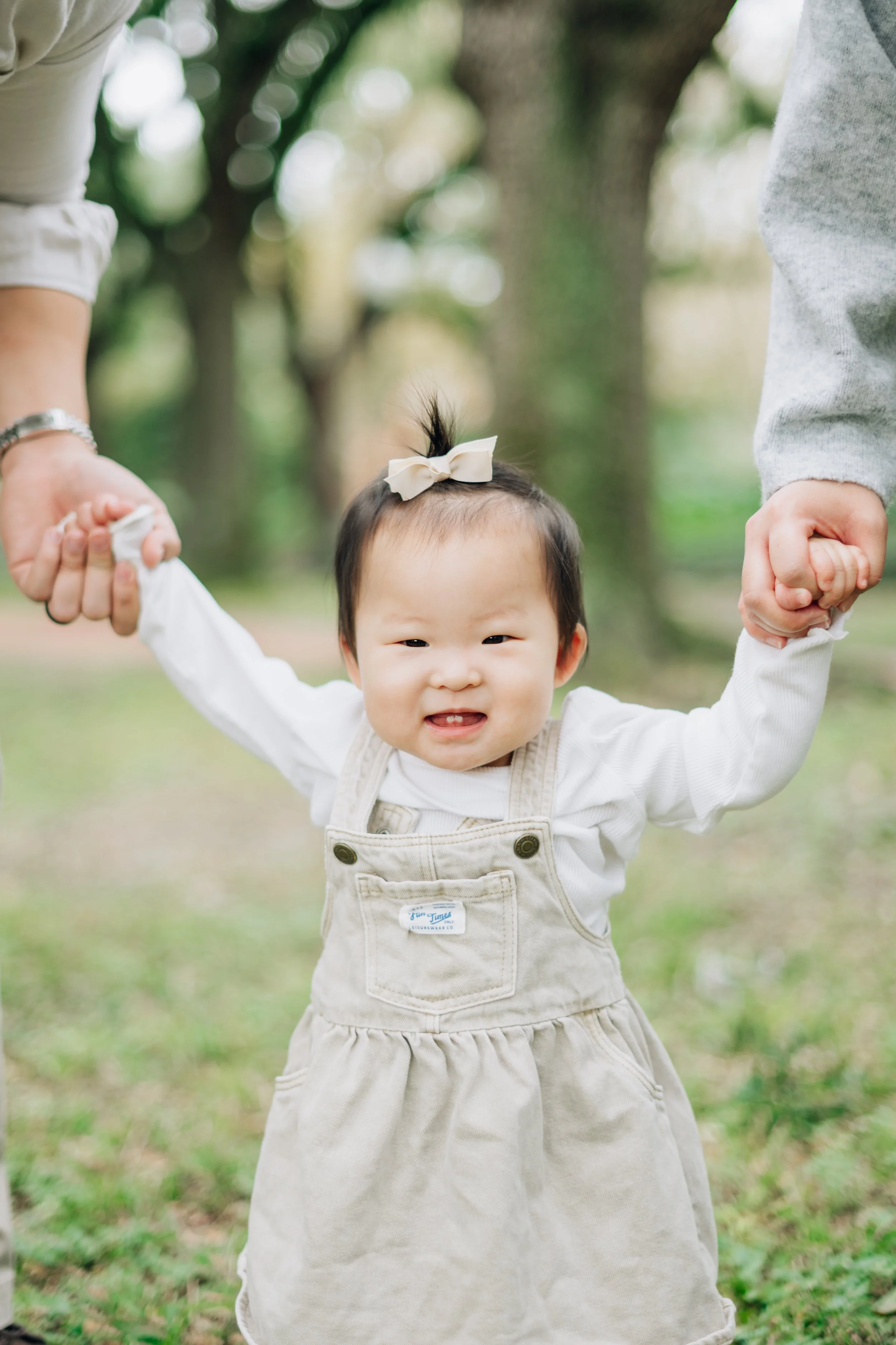 A happy young Asian girl with a bow in her hair, wearing beige overalls and a white shirt, holding hands with two adults in a park.