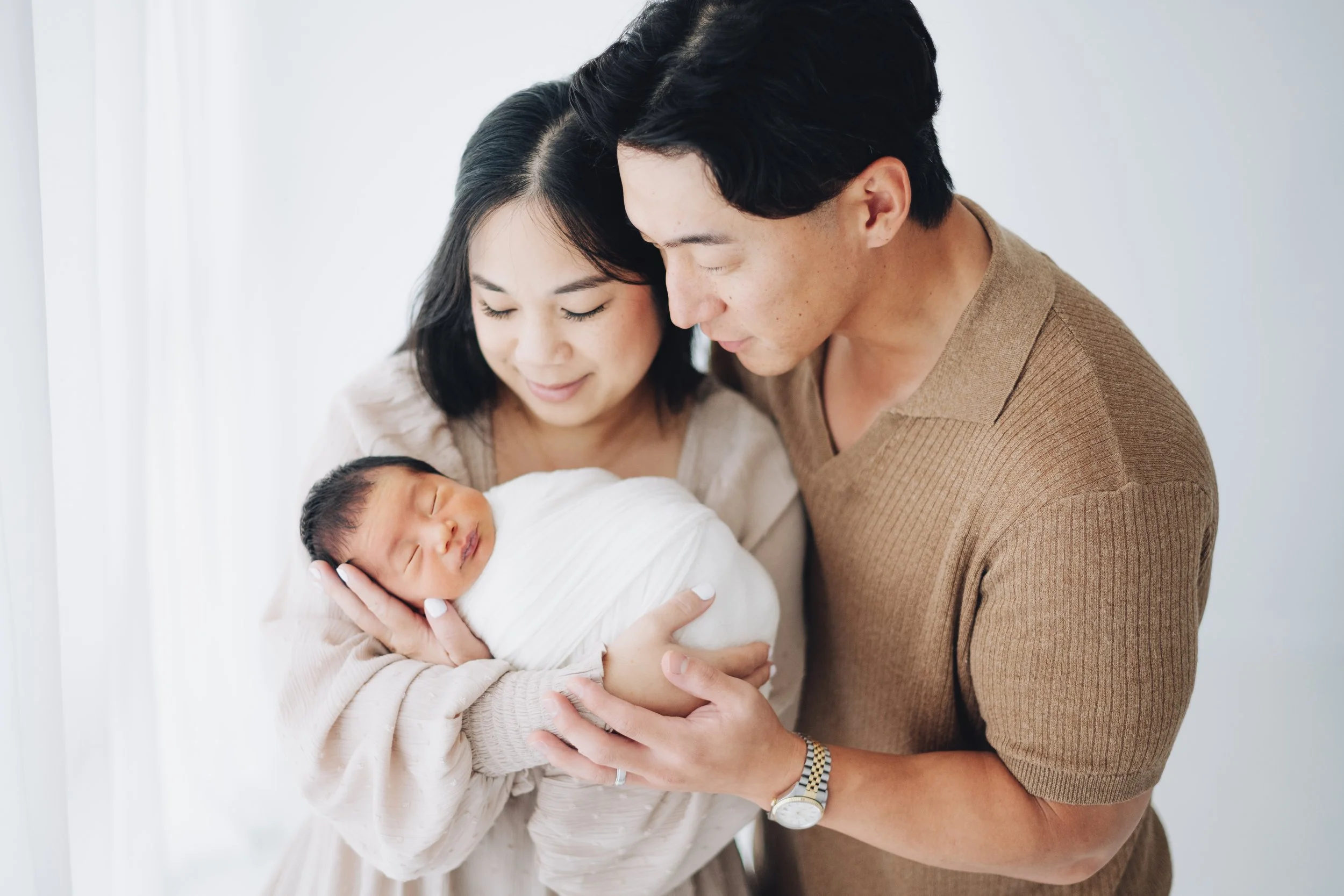 A couple holding a newborn baby wrapped in white swaddle, with the mother and father looking lovingly at the baby in a bright, minimalistic room.