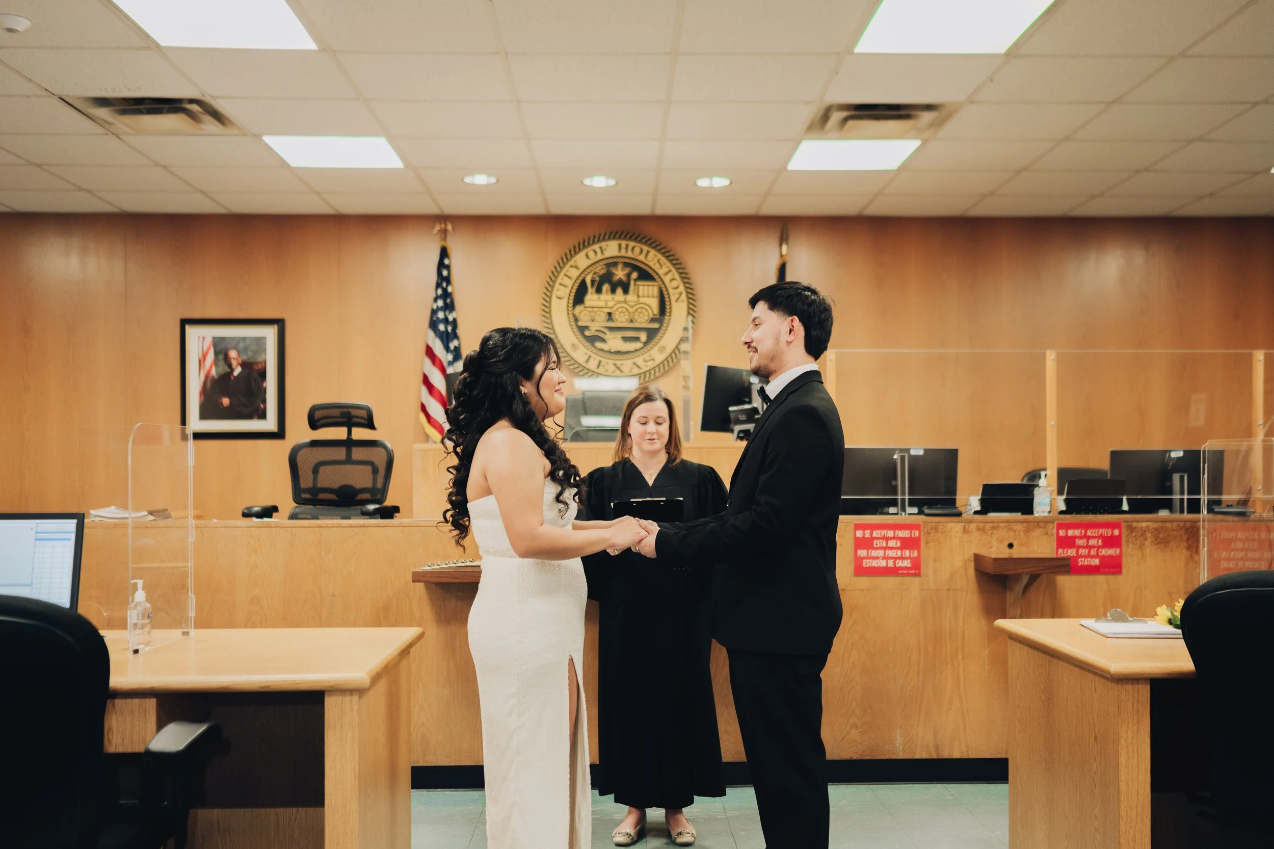 A bride and groom holding hands and looking at each other during their wedding ceremony in a city hall, with a judge presiding over the event and the Texas state flag and city seal visible in the background.