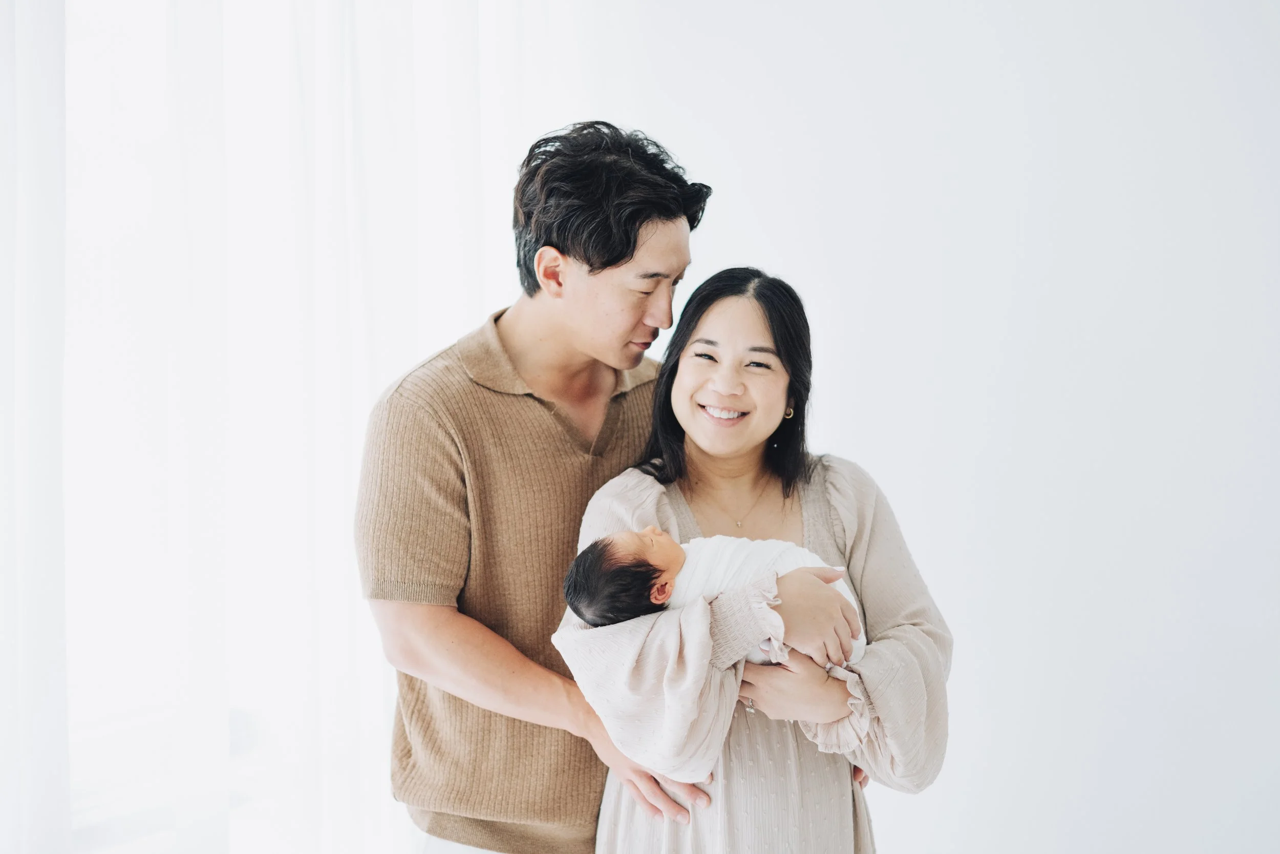 A happy woman holding a newborn baby wrapped in a white blanket, with a smiling man standing beside her in a bright, minimalistic room.