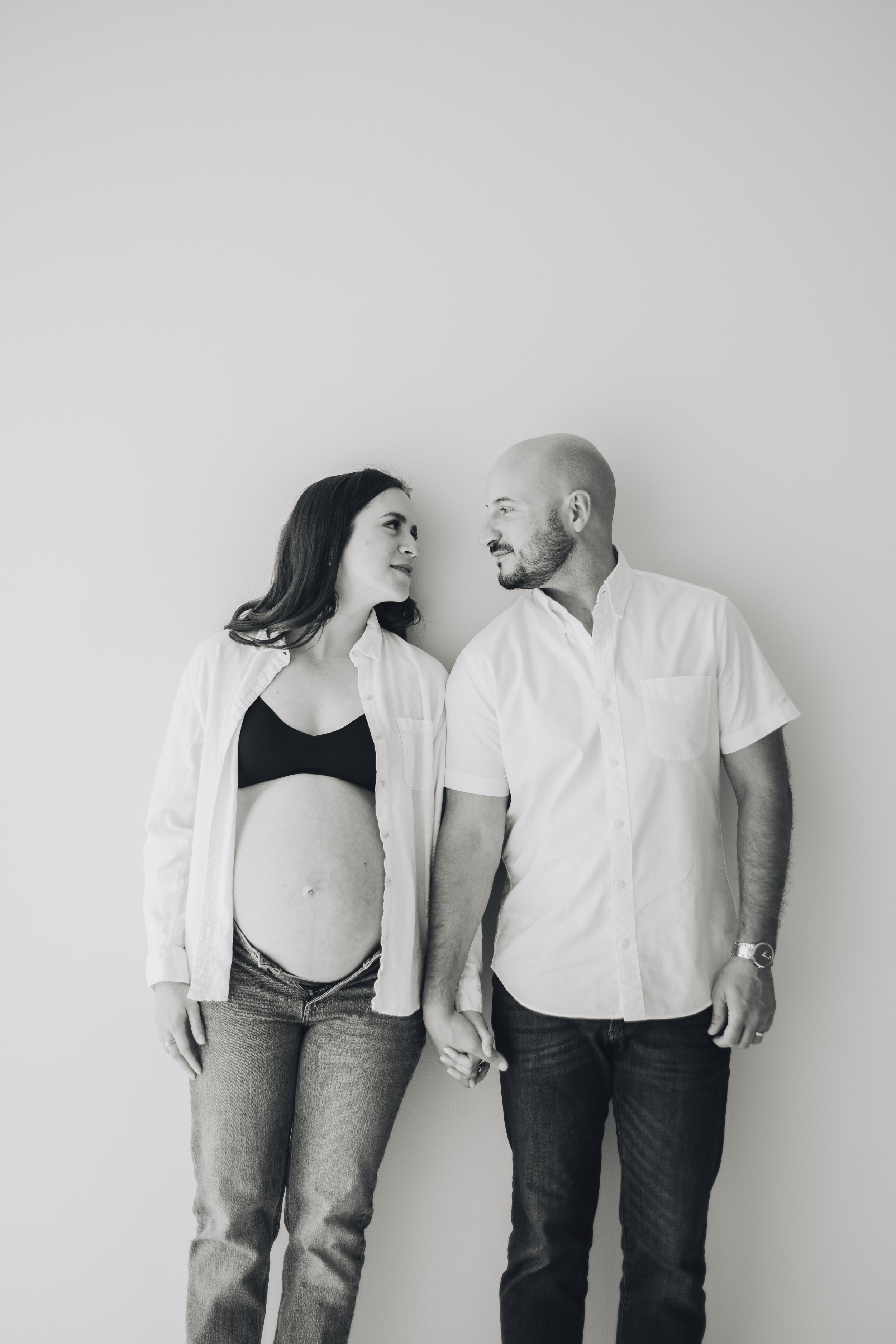 Pregnant woman and man holding hands, looking at each other, standing against plain wall.