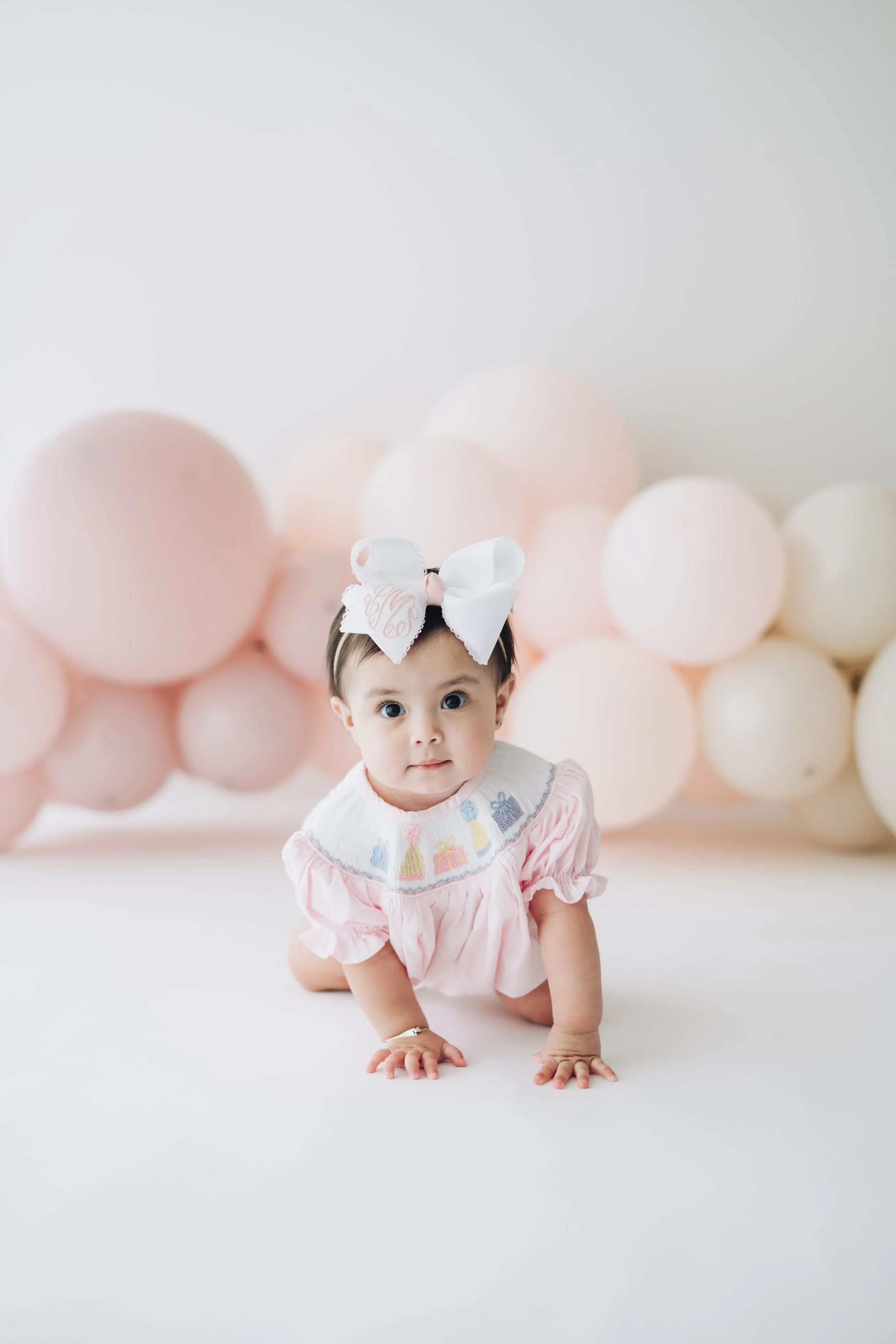 A baby girl crawling on a white floor, wearing a pink dress with embroidered collar and a large white bow headband, with pink and white balloons in the background.