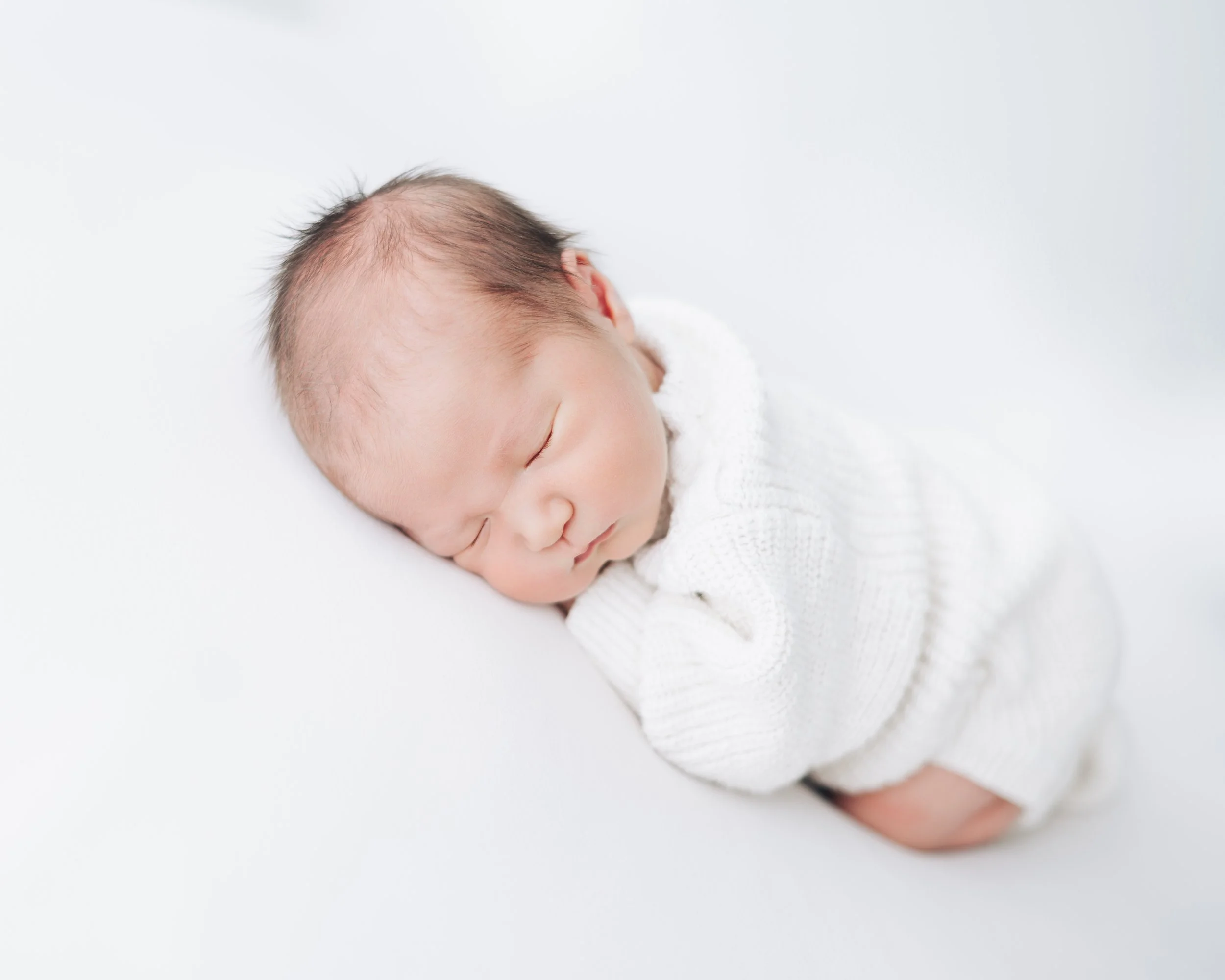 A sleeping baby wearing a white sweater, lying on a white surface.