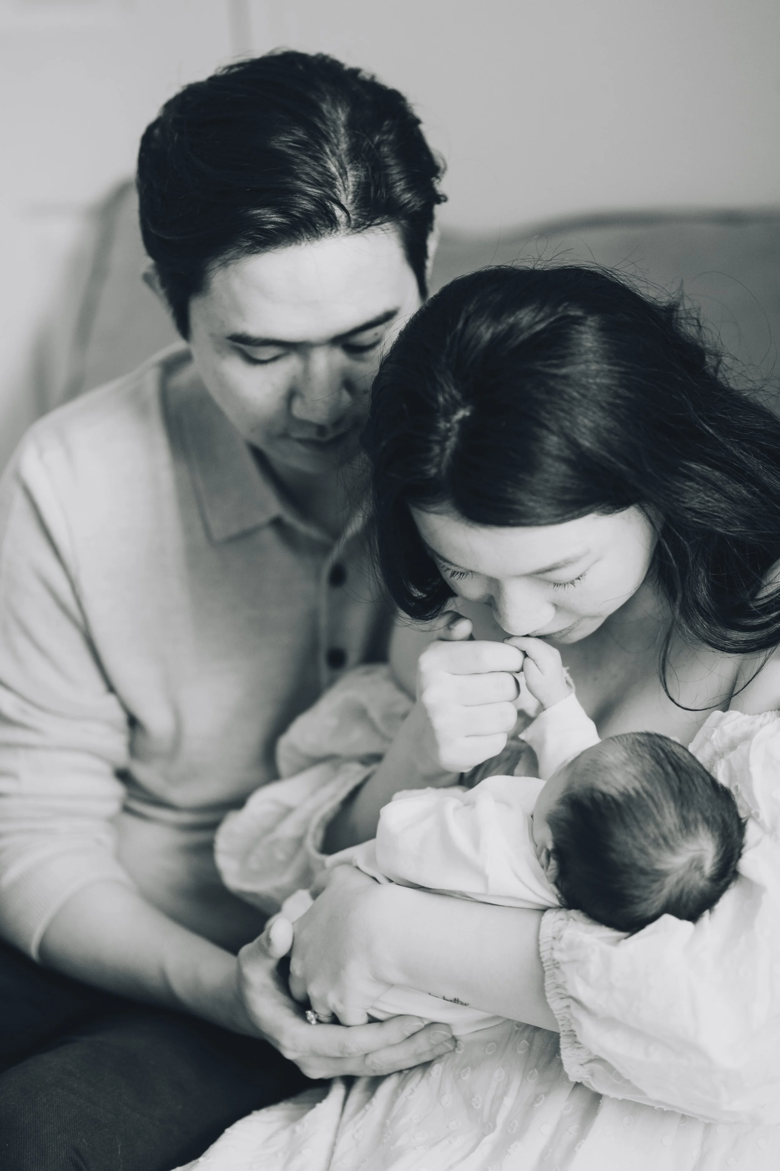 A black and white photograph of a family with a mother, father, and a newborn baby. The mother is holding the baby close to her chest, and the father is leaning in, both with gentle expressions of affection.