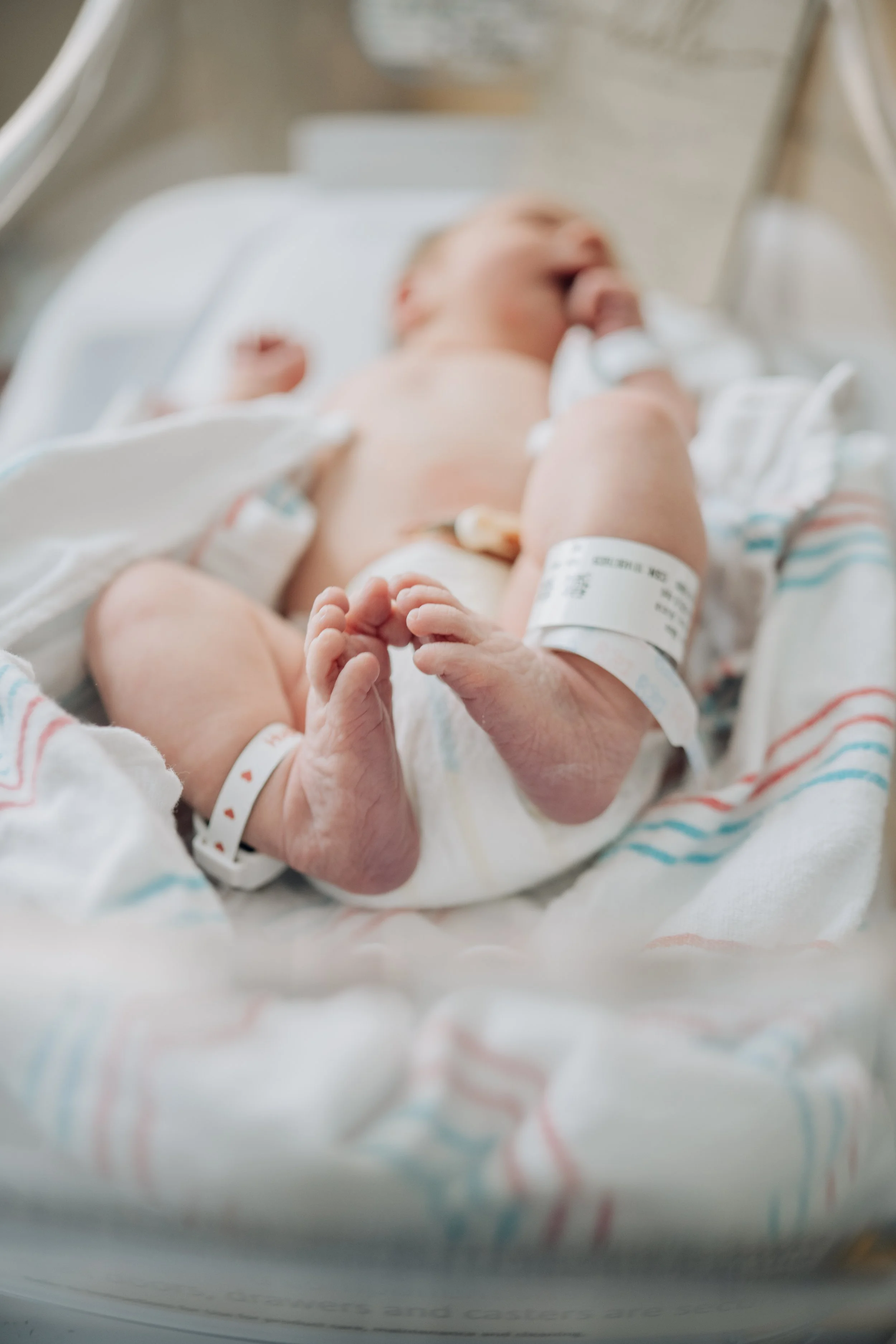 Newborn baby lying in a hospital bassinet, wrapped in a hospital blanket, with wristbands on both arms.
