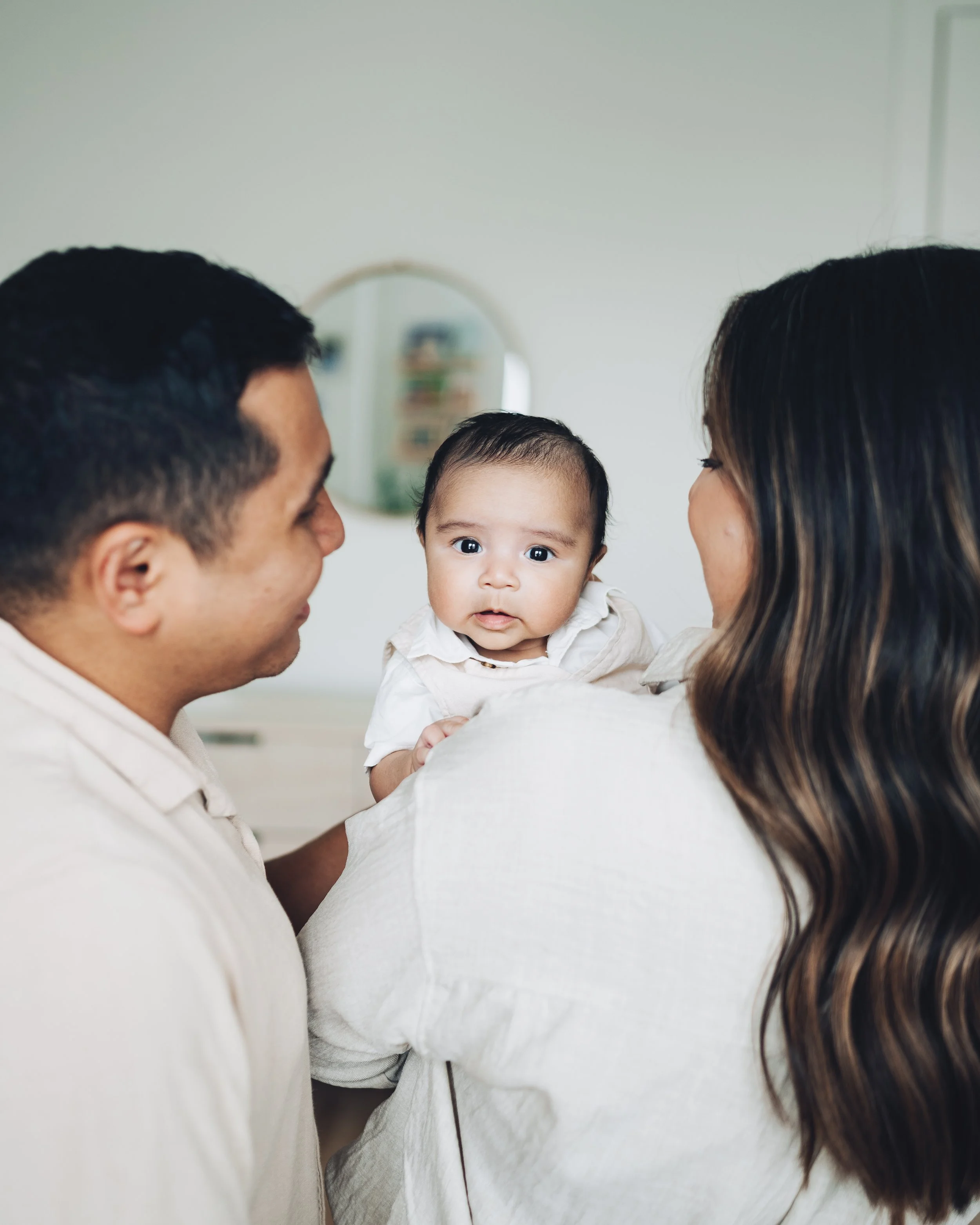 A family of three, a man and woman holding a baby, looking at each other in a bright room with a mirror in the background.