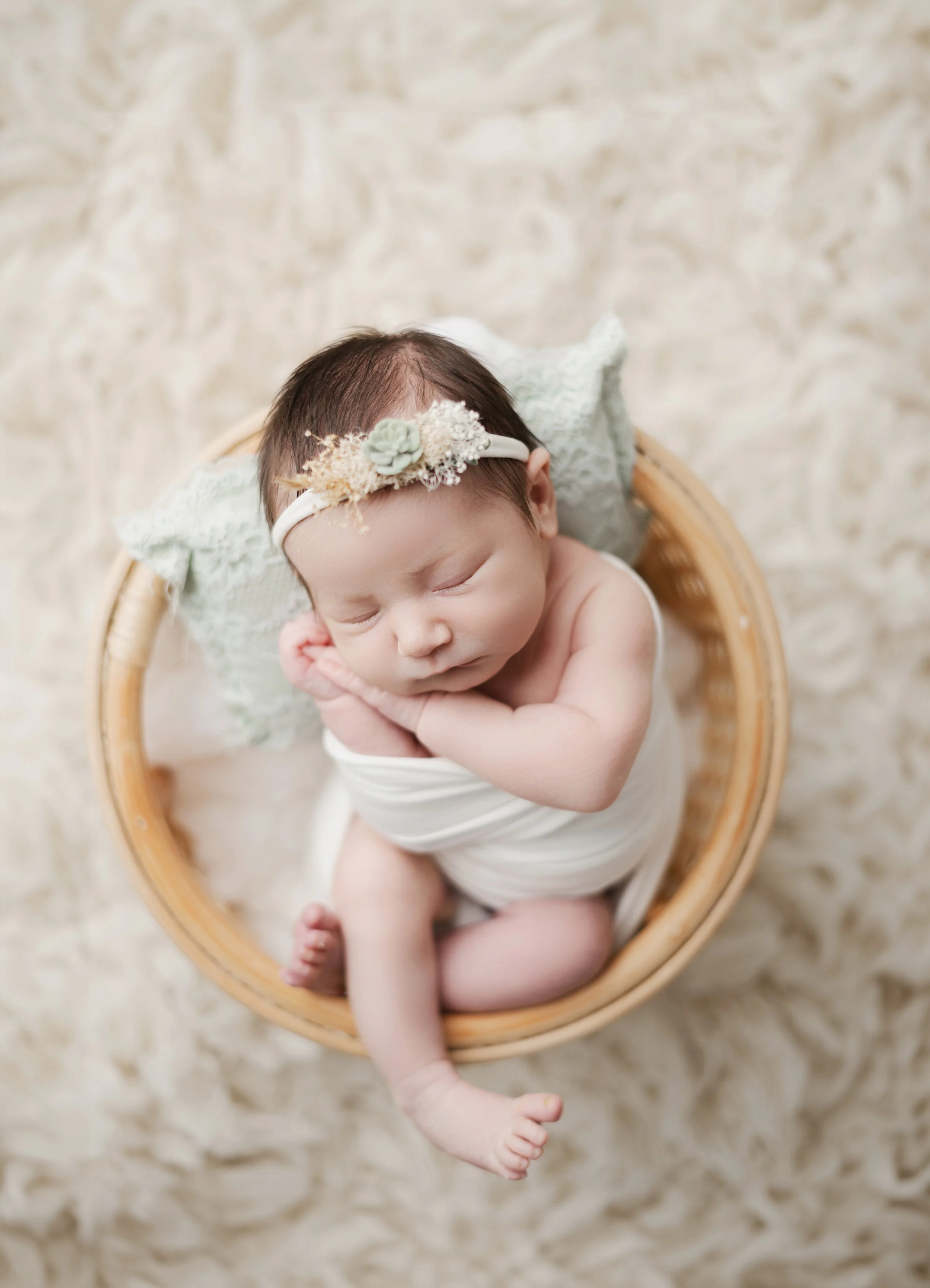 A sleeping newborn baby in a woven basket, wrapped in white cloth, wearing a floral headband, on a soft cream-colored blanket.