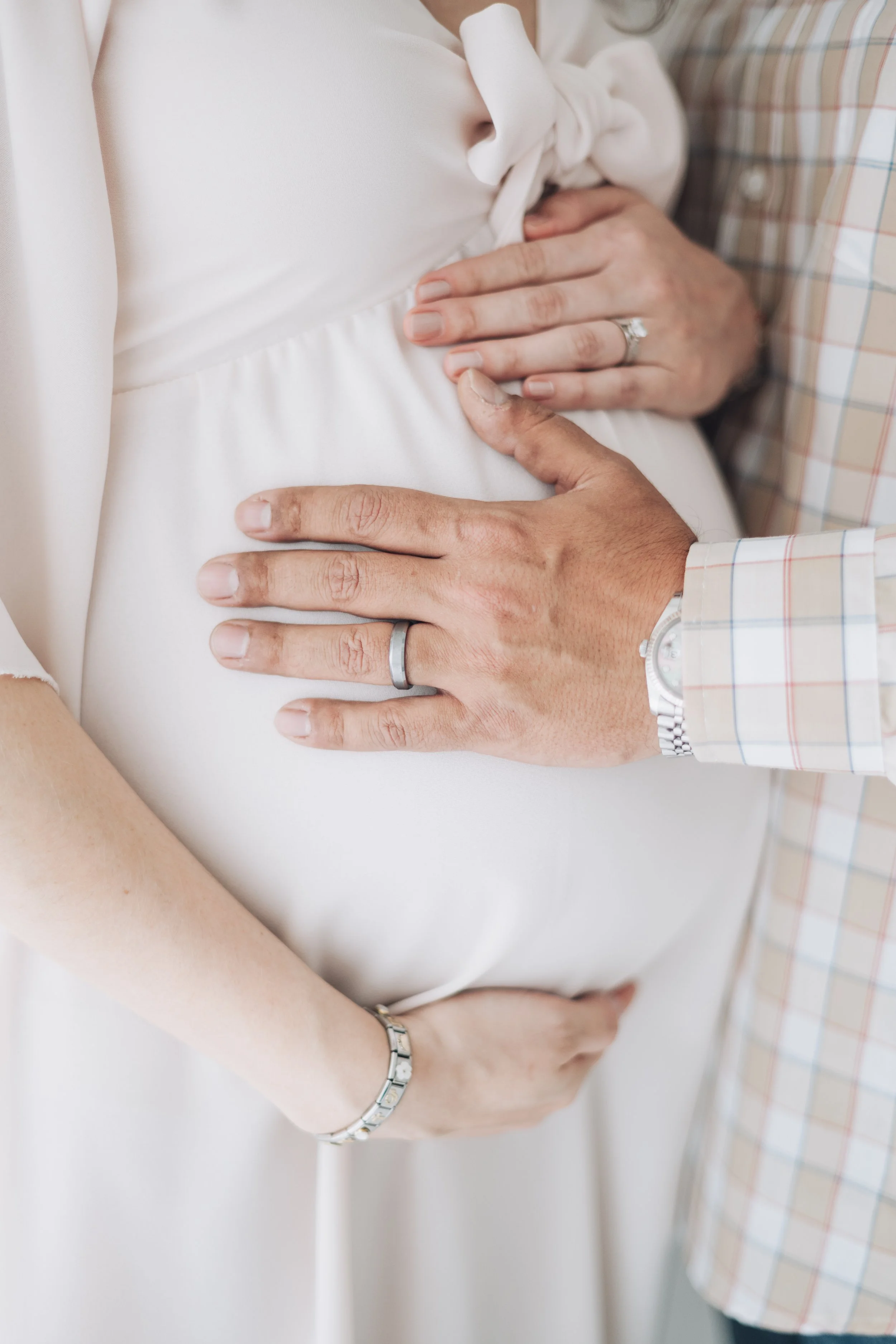 Close-up of a pregnant woman with her partner's hands gently resting on her belly, showing wedding rings on their ring fingers.