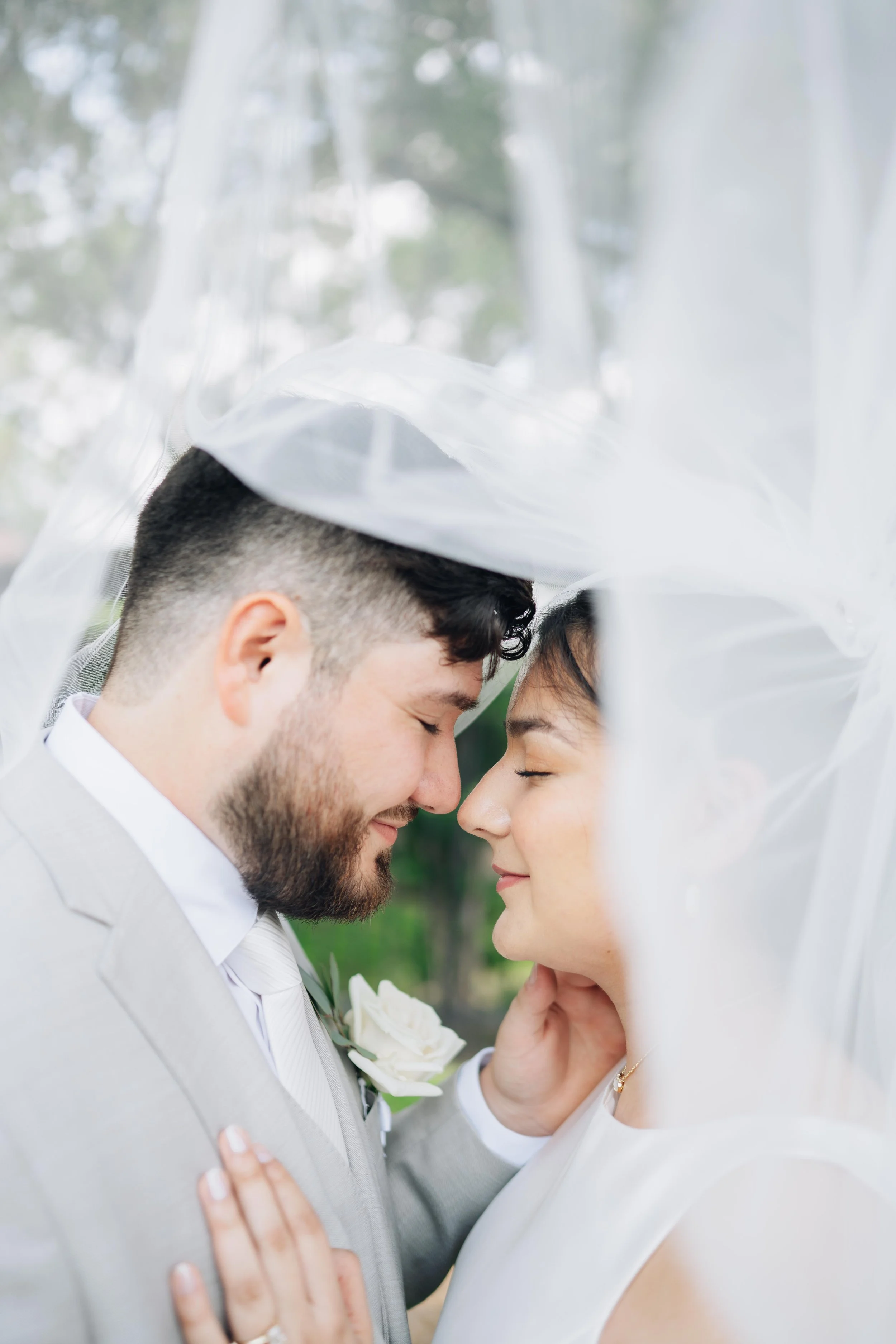 A bride and groom with foreheads touching, eyes closed, under a sheer veil outdoors during their wedding. The groom is wearing a light gray suit with a white shirt and a white rose boutonniere, while the bride is in a white dress with a necklace.