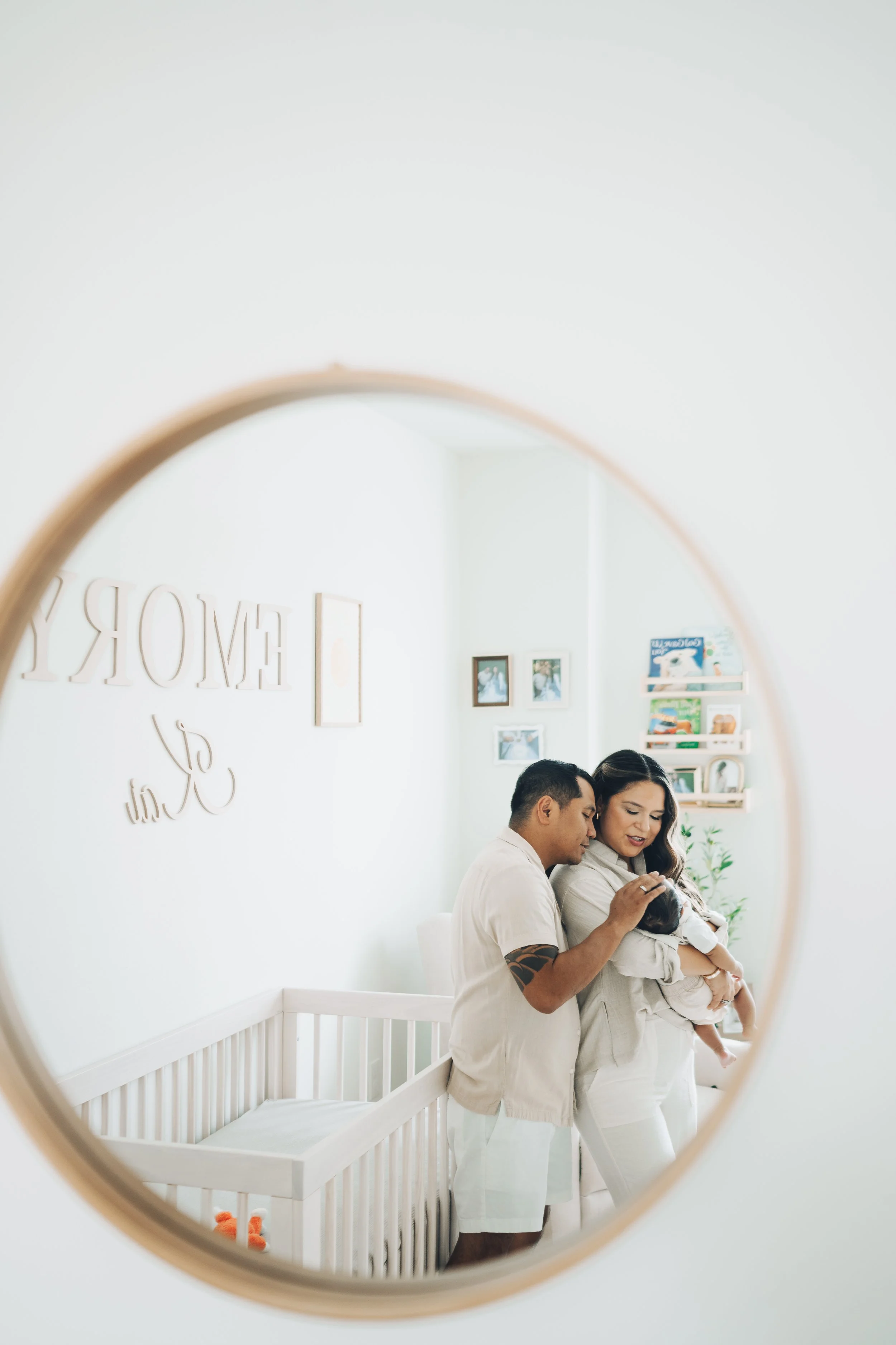 A couple looking into a mirror in a baby's nursery, with a woman holding a baby.