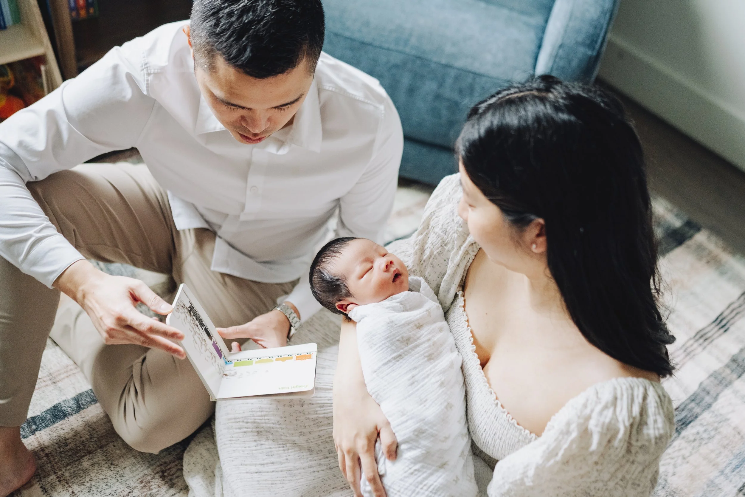 A mother holding a newborn baby, with a man showing a photo on a tablet, in a cozy living room.