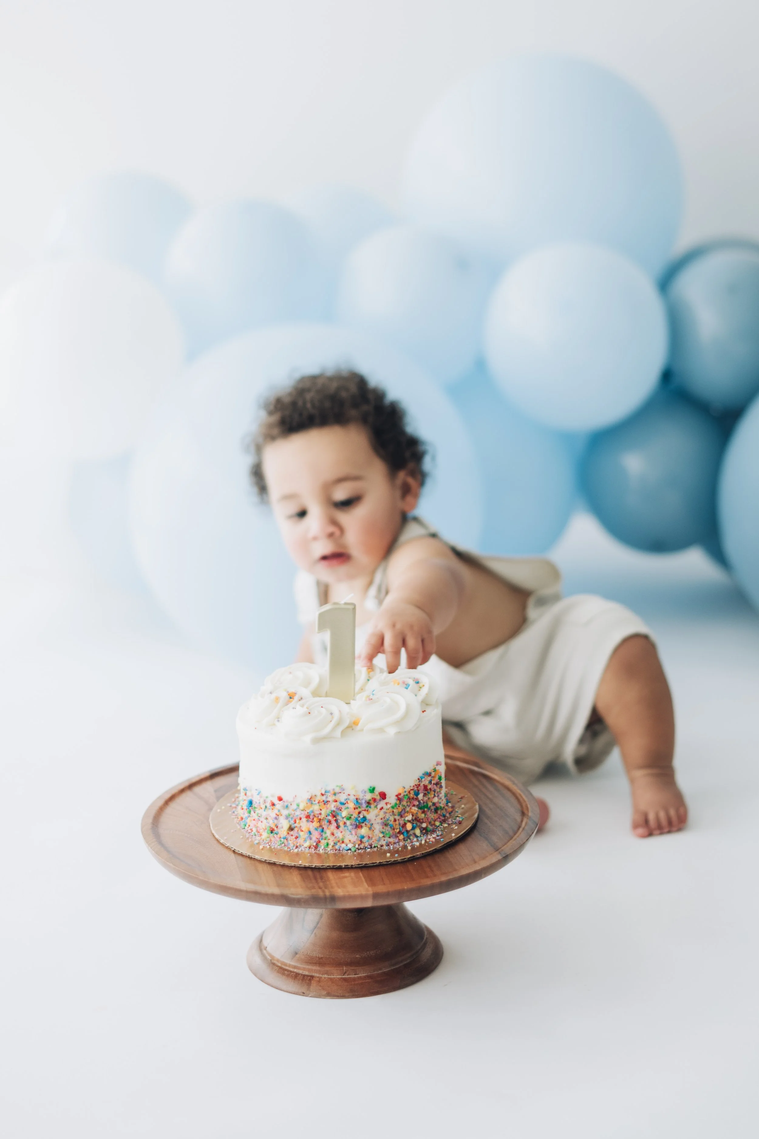 Young child reaching for a birthday cake decorated with sprinkles and a candle shaped as the number 1, with blue balloons in the background.