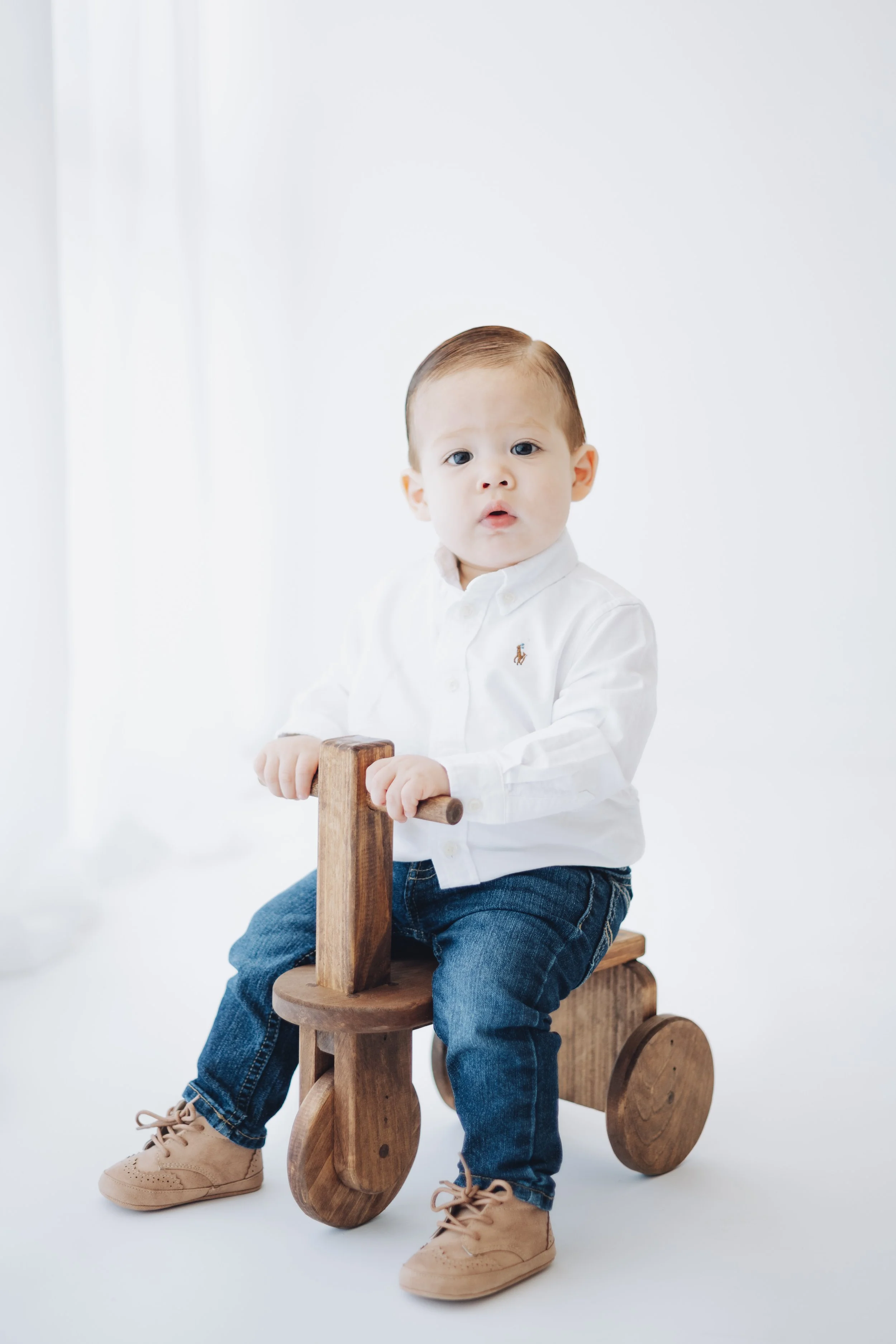 A young boy with light skin, wearing a white shirt, blue jeans, and tan shoes, sitting on a wooden ride-on toy against a plain white background, looking at the camera.