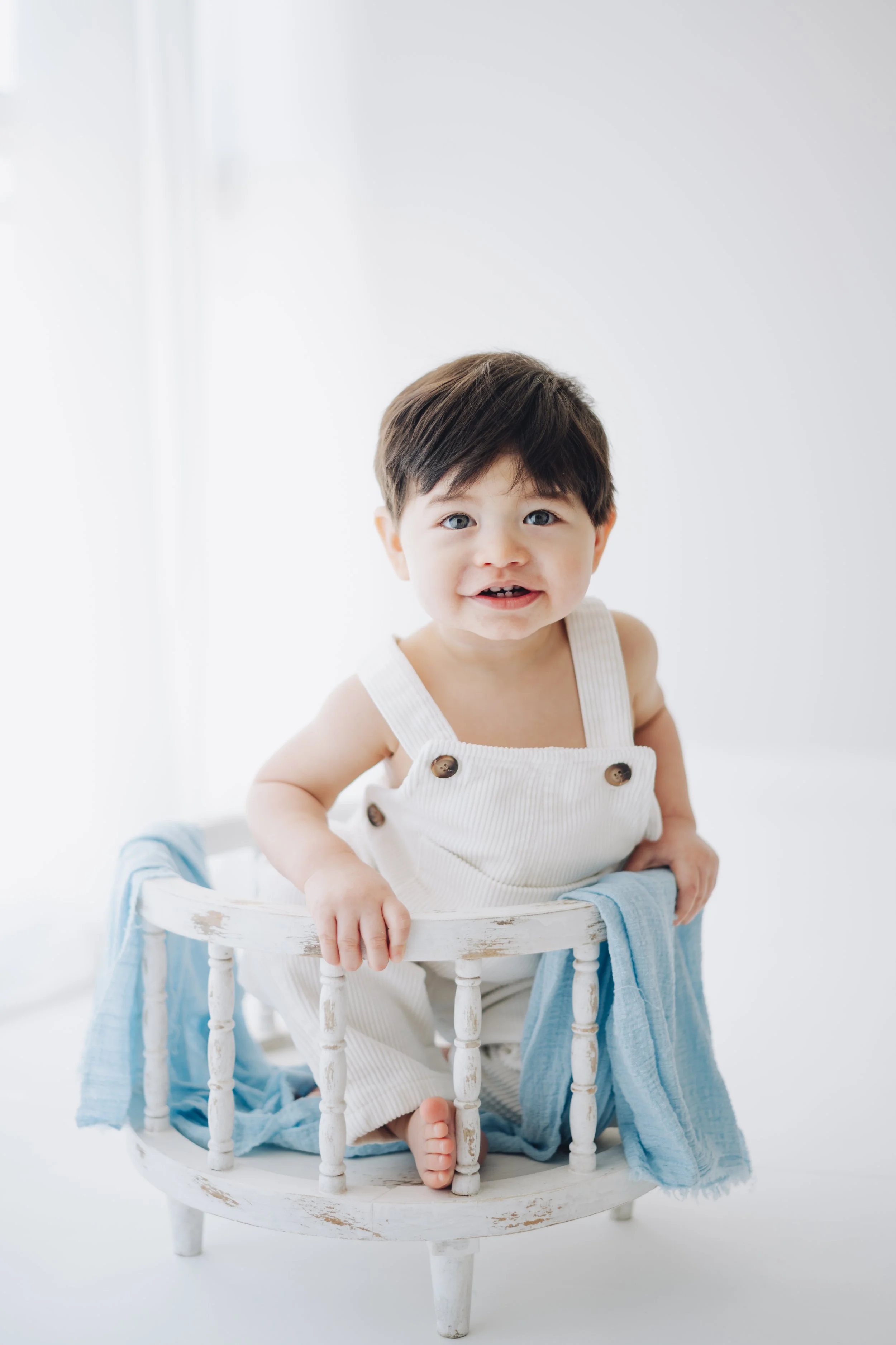 Smiling toddler holding onto a small white distressed wooden chair with a blue cloth draped over it, in a bright room.