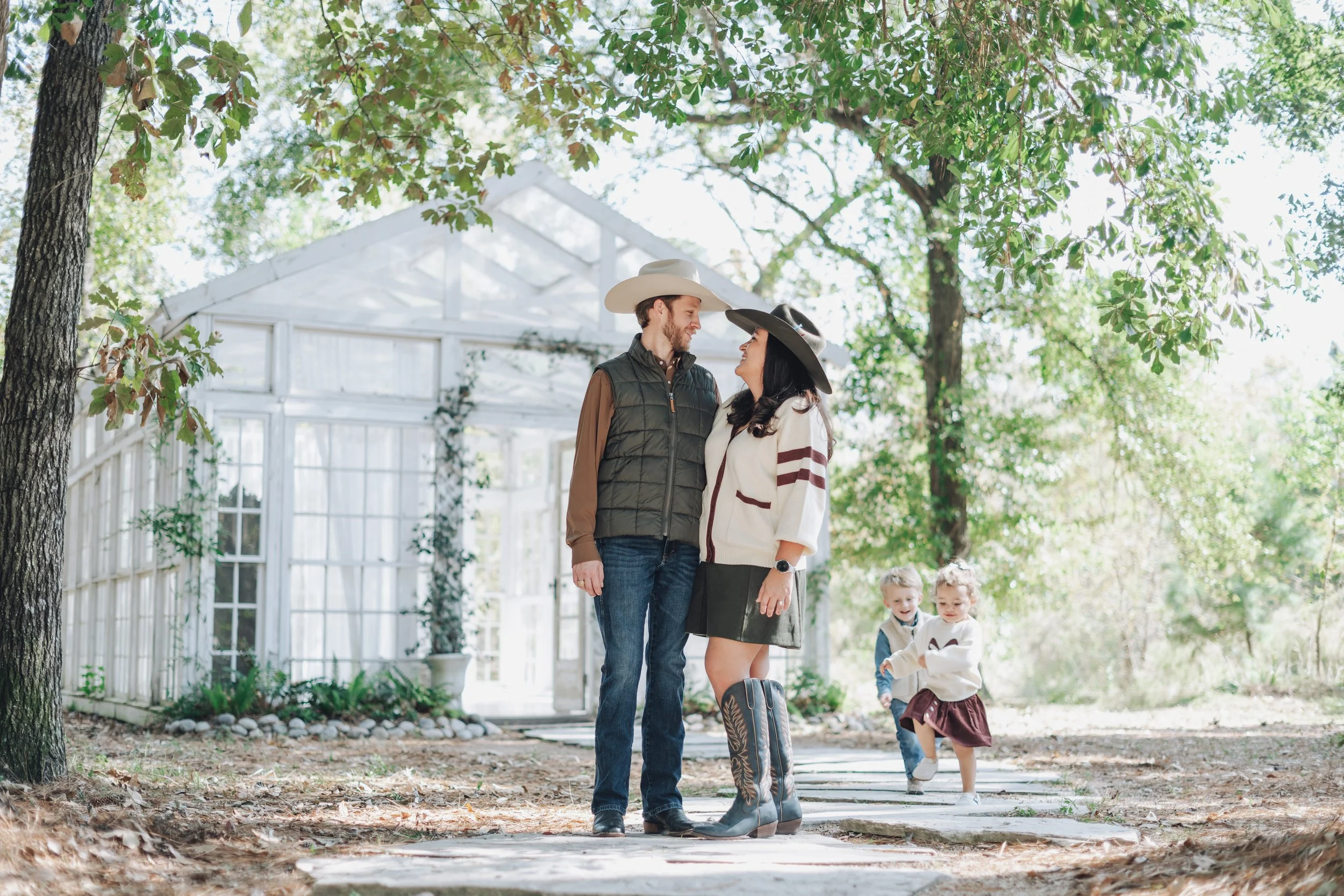 A couple standing in front of a greenhouse outdoors, smiling at each other, with two children playing on a stone pathway behind them.