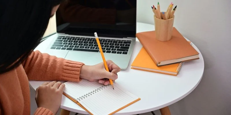 A woma writing in a notebook with her computer  by her.