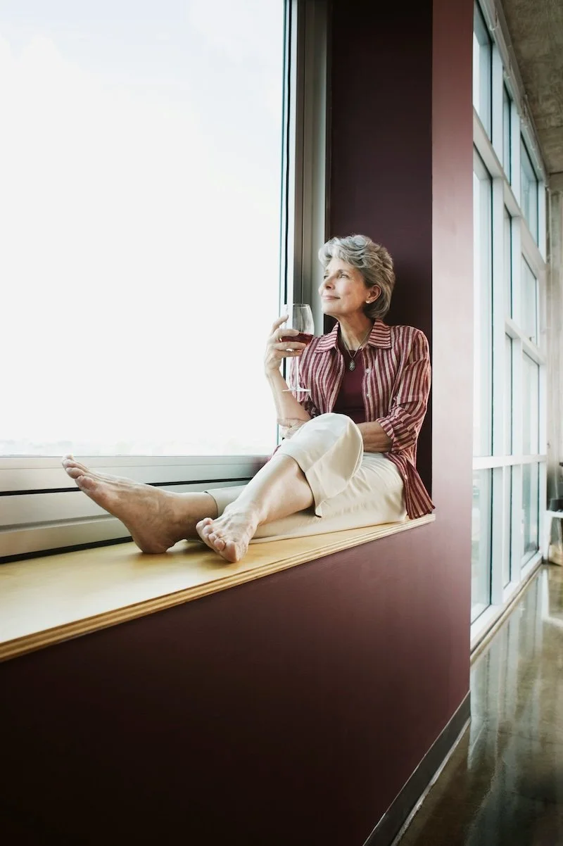 A woman over 55 sitting on a windowsill enjoying a drink of wine