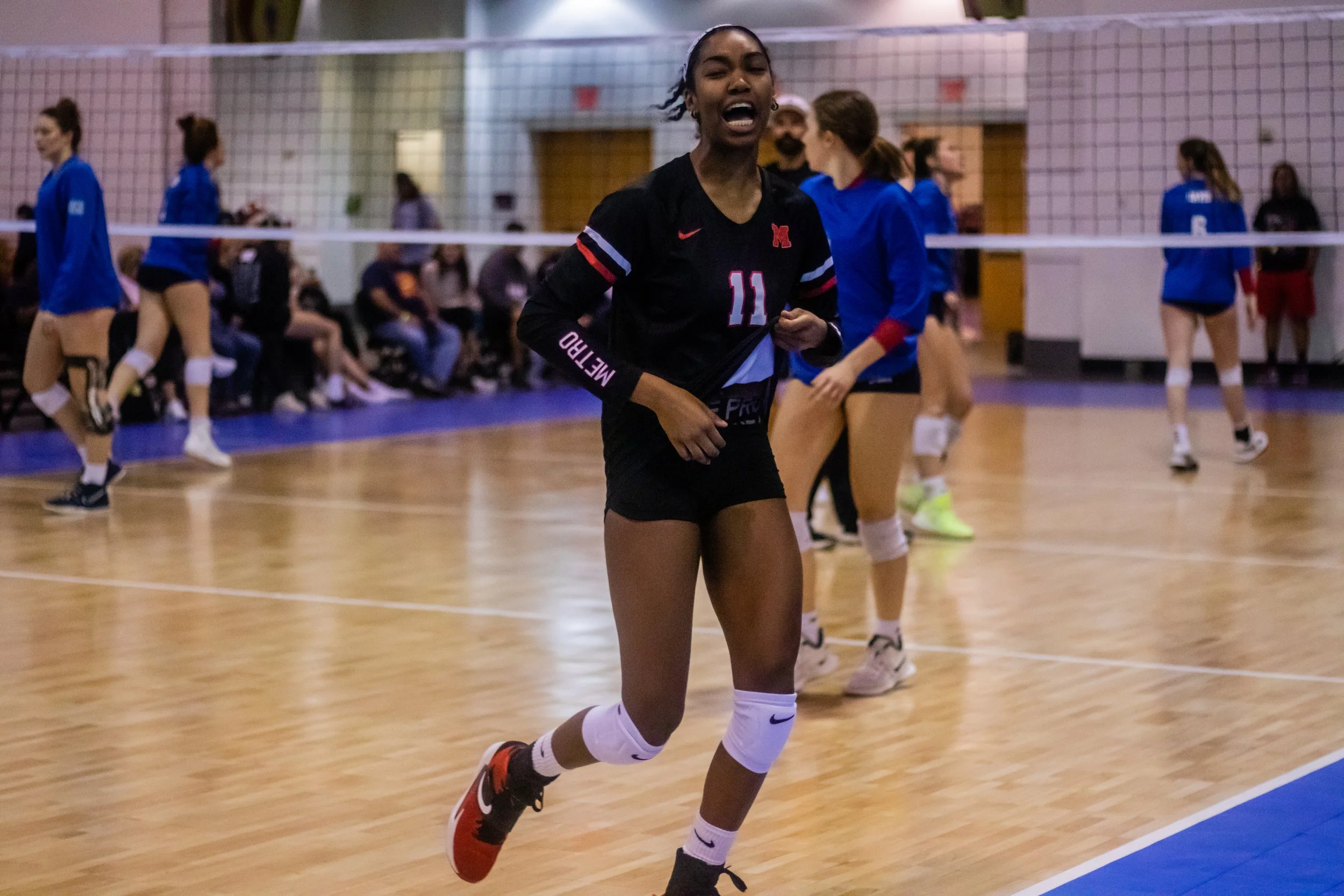 A female volleyball player in a black uniform with the number 11 is running on the court during a game, with other players and spectators in the background.