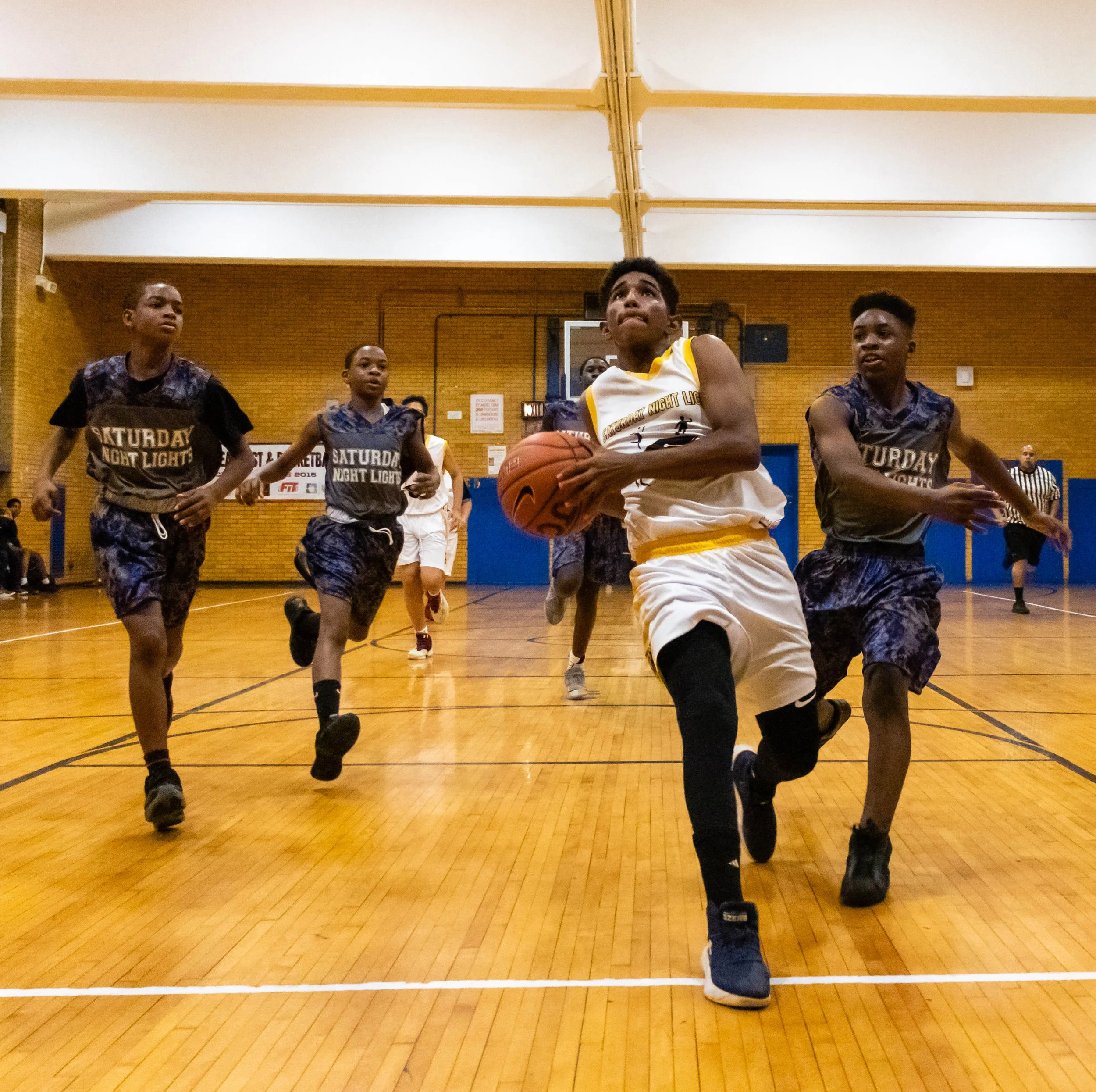 Young boys playing basketball in an indoor court, with one boy in a white jersey holding the ball and running towards the basket, surrounded by other players in dark jerseys.